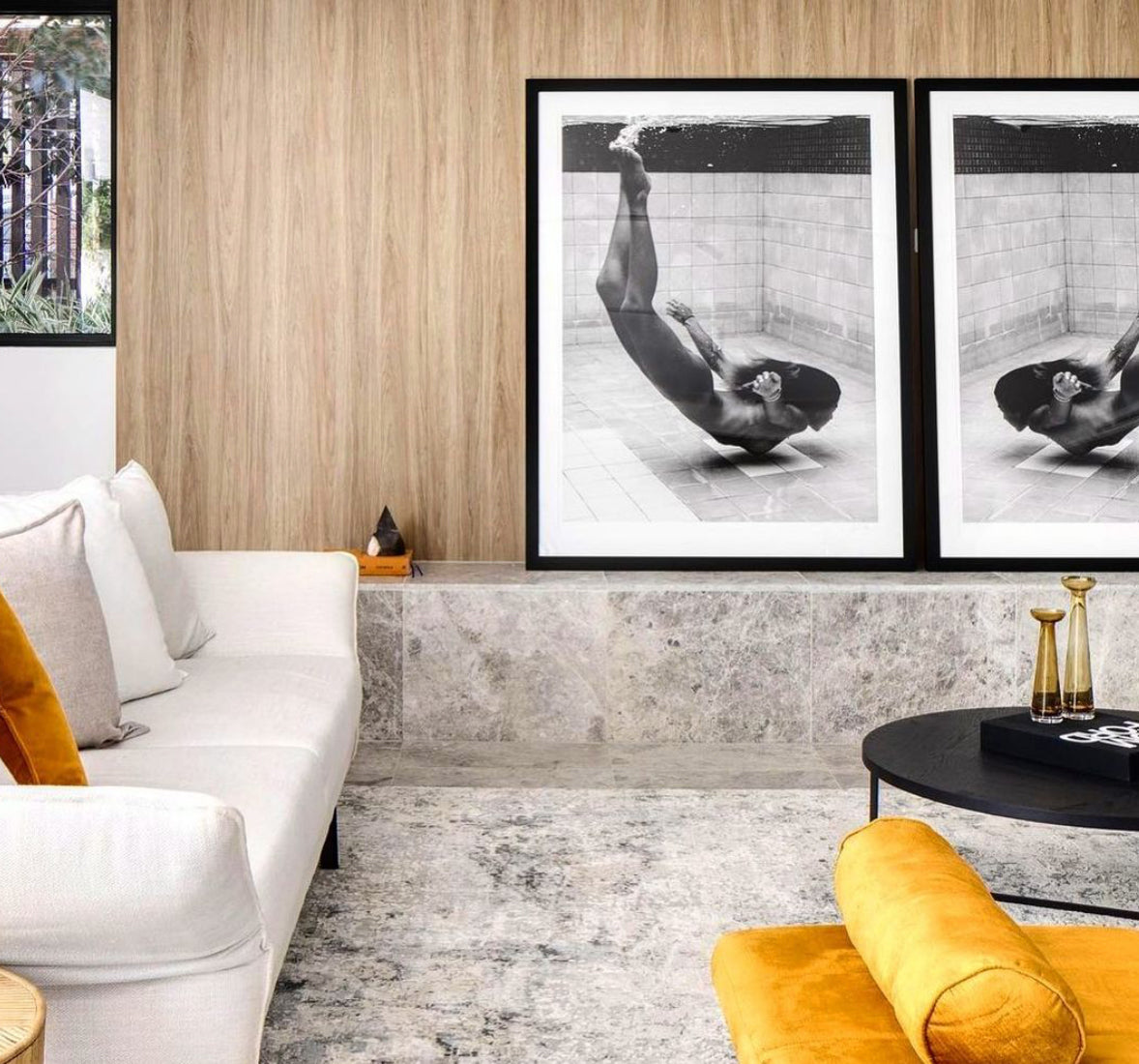 A modern living room featuring a white sofa with beige and yellow cushions, a circular black coffee table with gold accents, and a yellow armchair. The wooden wall behind the sofa displays two large black and white art prints titled "Fall" by Francesca Owen, elegantly showcased with custom framing.