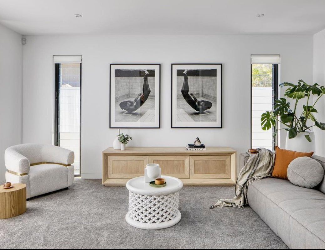 A modern living room features a light gray carpet and a gray sofa adorned with orange and white pillows, centered around the "Fall" round white coffee table by Francesca Owen. A matching white armchair sits nearby. The walls are decorated with two black-and-white art prints on fine art paper, complemented by custom framing. The space is brightened by natural light and includes a large potted plant in the corner.