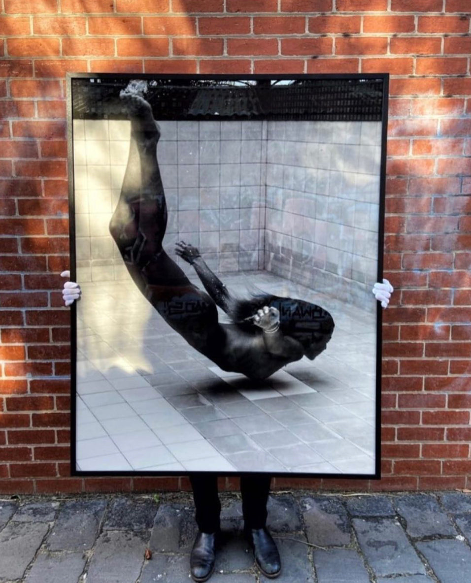 A person wearing black shoes and white gloves holds up Francesca Owen's "Fall" in front of them. The large framed photograph features a striking black and white image of a diver mid-dive, creating the illusion that the diver's legs extend out of the frame. The artwork is printed on fine art paper and set against a brick wall backdrop.