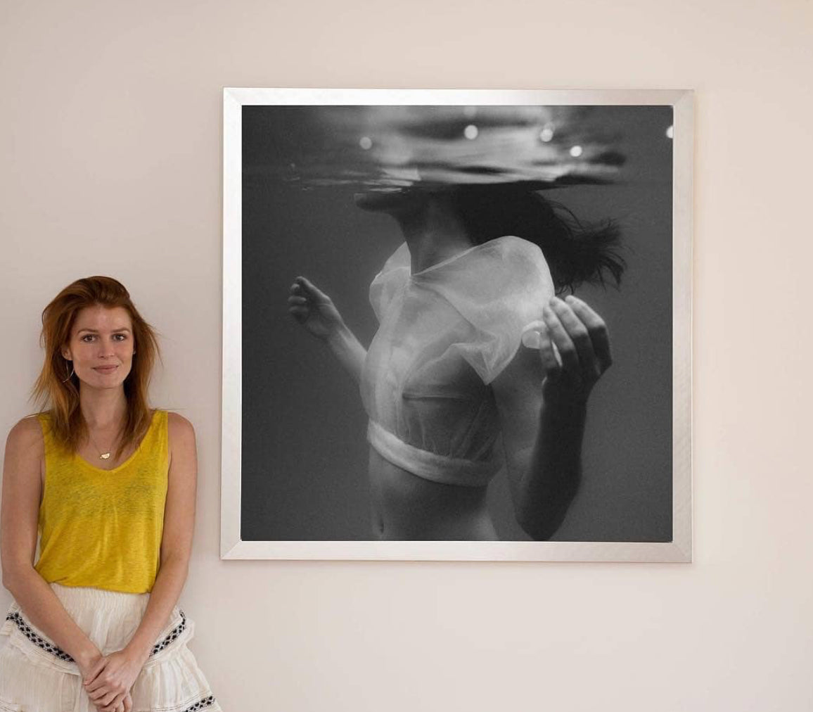 A woman with auburn hair, dressed in a yellow sleeveless top and a white skirt, stands smiling next to "Accepting," a framed black-and-white photograph by Francesca Owen. The fine art paper print features a submerged person wearing a translucent top, their head tilted back and arms outstretched. It utilizes archival pigments to ensure lasting quality.
