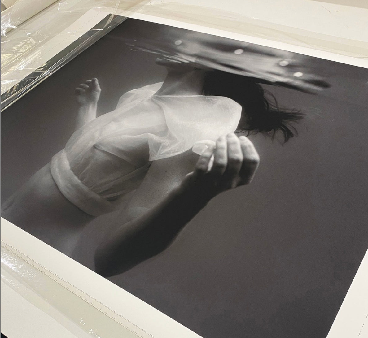 Accepting" by Francesca Owen is a compelling black and white photograph showcasing an underwater scene. In the image, a person in a sheer top can be seen with their head partially submerged and their body ascending towards the surface. This artwork is printed using archival pigments on fine art paper, framed or mounted flat, and partially covered with plastic wrap.