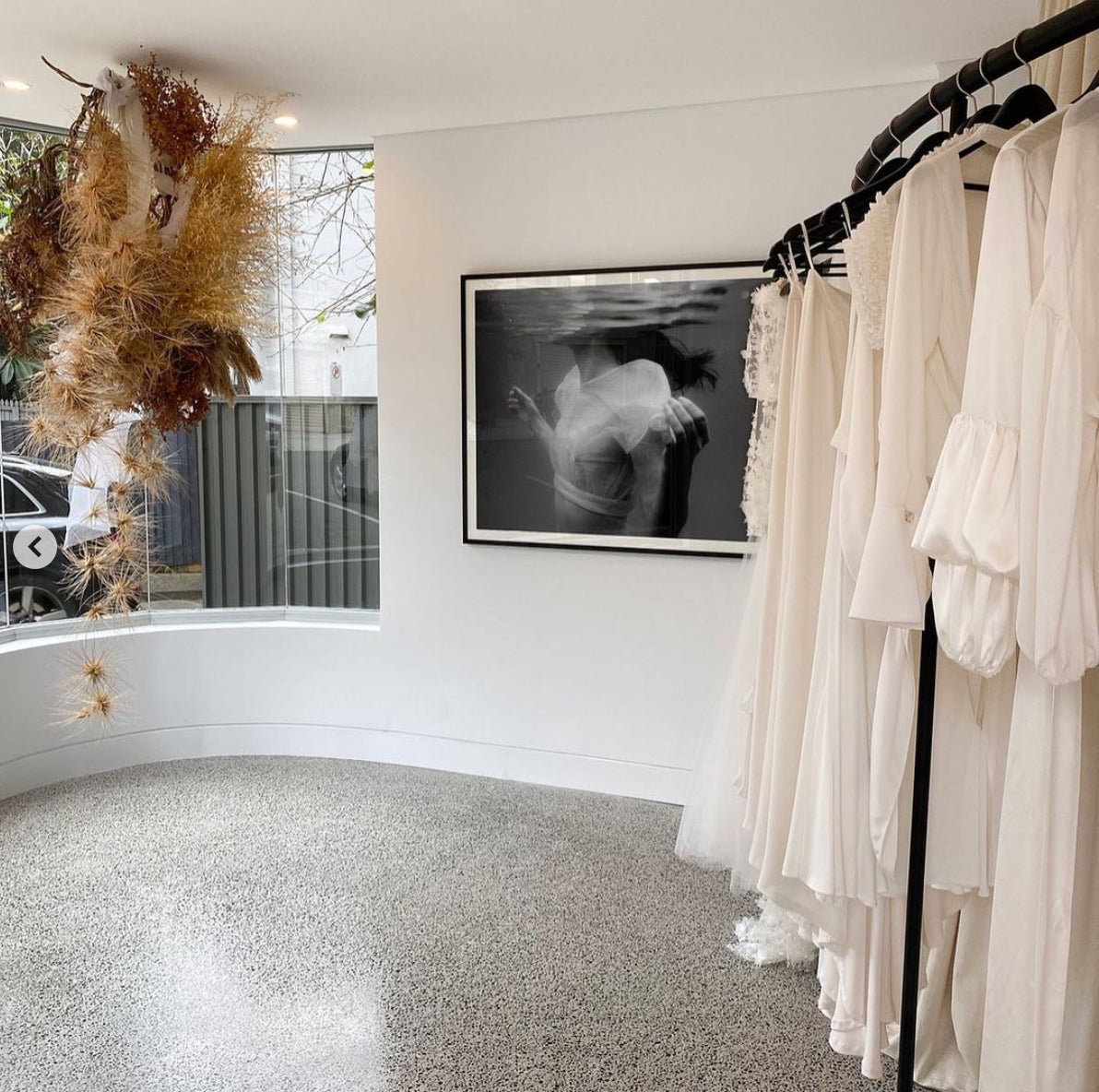 A minimalist bridal boutique features a rack of white wedding dresses on the right side, "Accepting," a black-and-white photograph by Francesca Owen of a couple underwater printed on fine art paper with archival pigments, and a large arrangement of dried flowers hanging from the ceiling by a curved window.