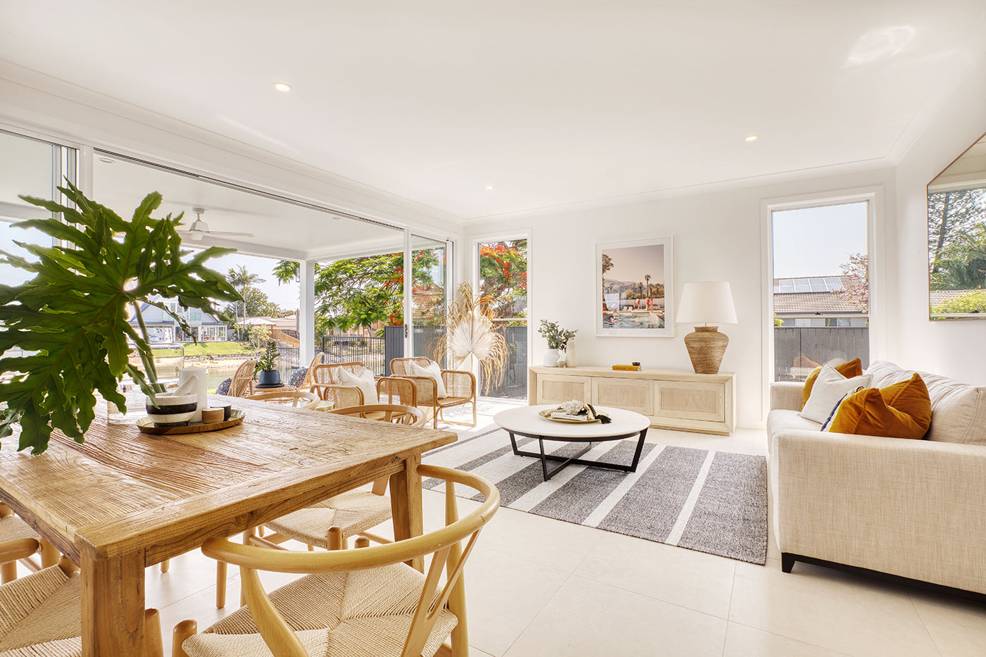 A bright, modern living room with large windows features a beige sofa with orange cushions, a wooden coffee table on a striped rug, and a wooden dining table with matching chairs. Adorning the walls are limited edition prints of Francesca Owen’s "Poolside Mermaids" on fine art paper, each accompanied by a certificate of authenticity.