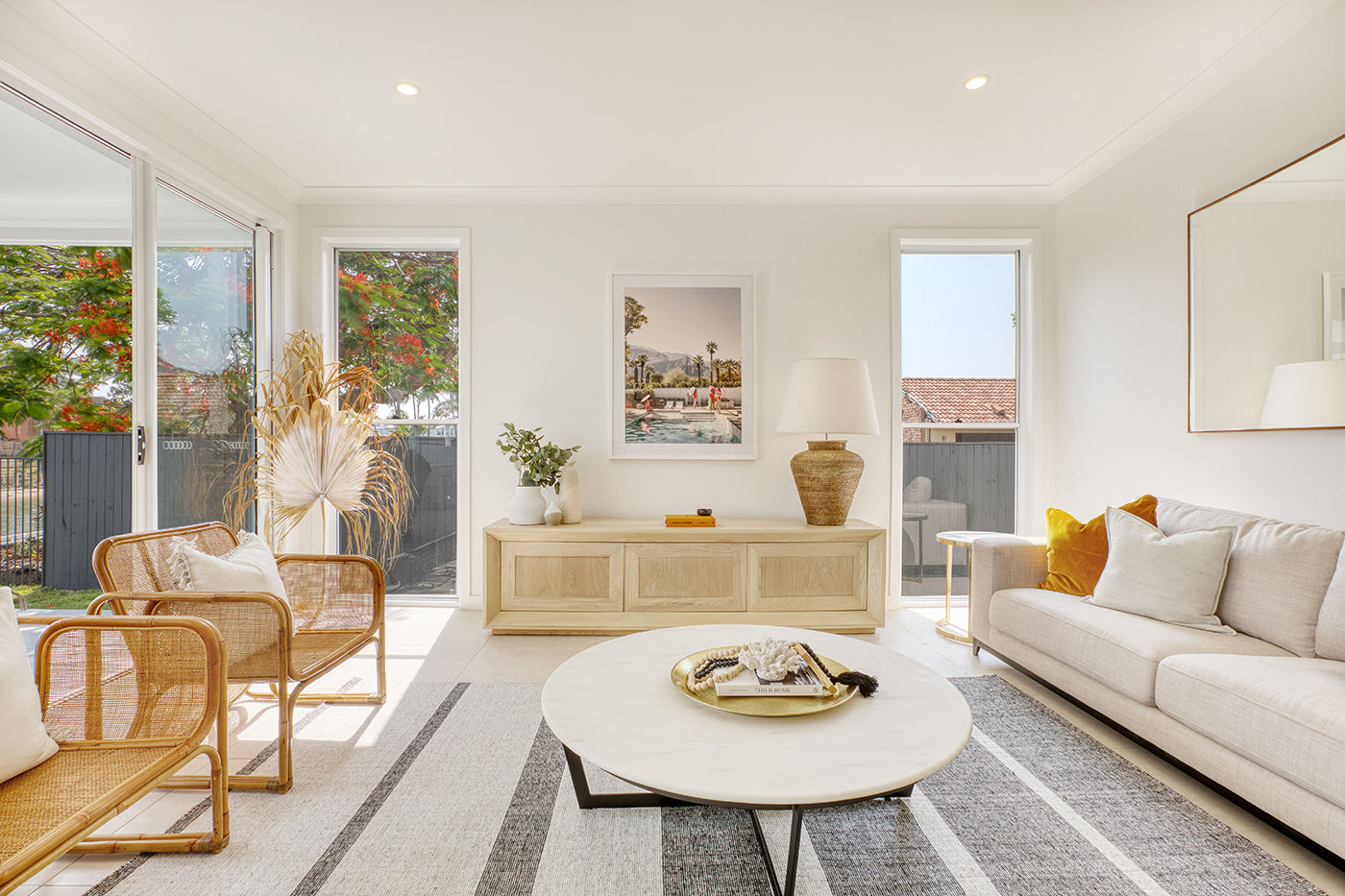 A bright, modern living room with large windows features a beige sofa with orange cushions, a wooden coffee table on a striped rug, and a wooden dining table with matching chairs. Adorning the walls are limited edition prints of Francesca Owen’s "Poolside Mermaids" on fine art paper, each accompanied by a certificate of authenticity.