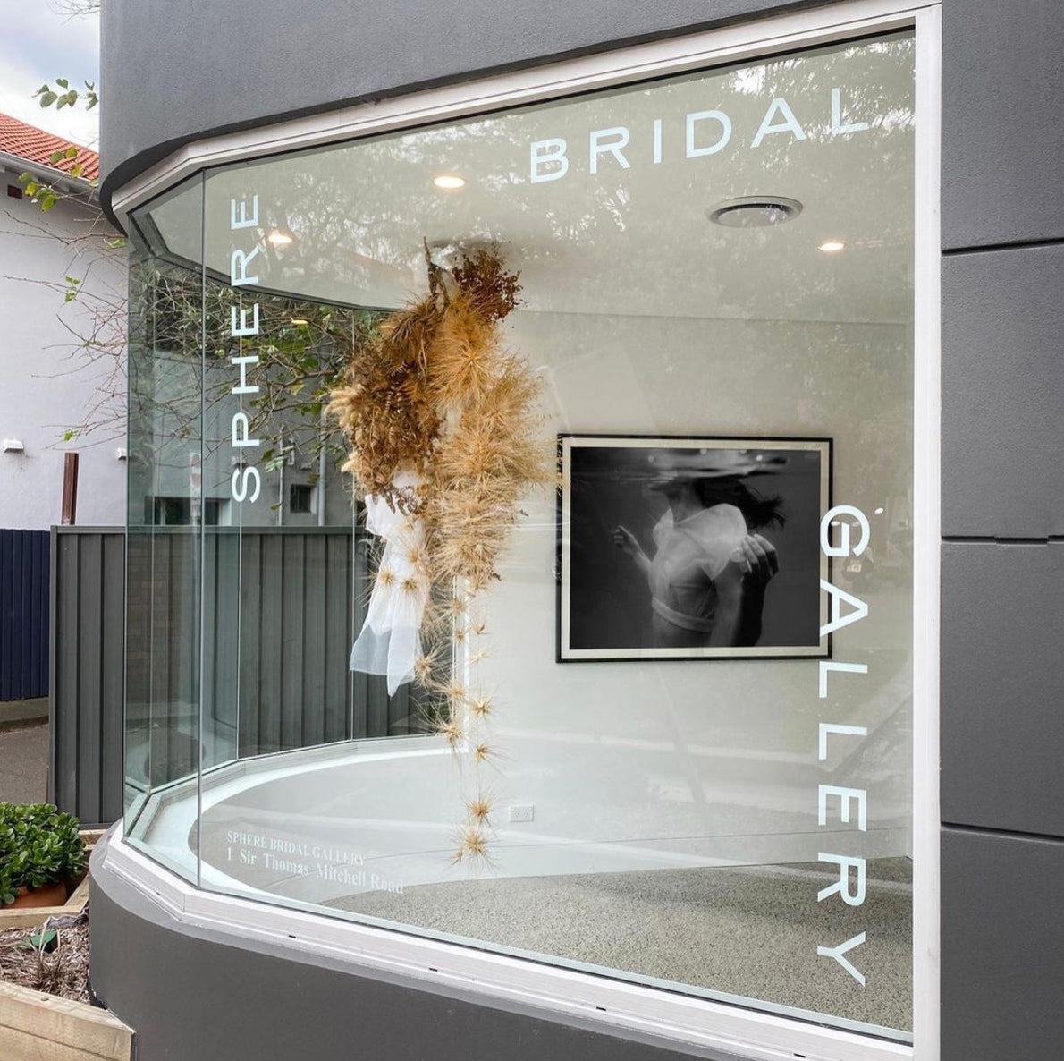 The shop window labeled "SPHERE BRIDAL GALLERY" showcases a display featuring dried floral arrangements and "Accepting," an elegant black-and-white photograph by Francesca Owen of an embracing couple printed on fine art paper with archival pigments for longevity, accompanied by a certificate of authenticity.