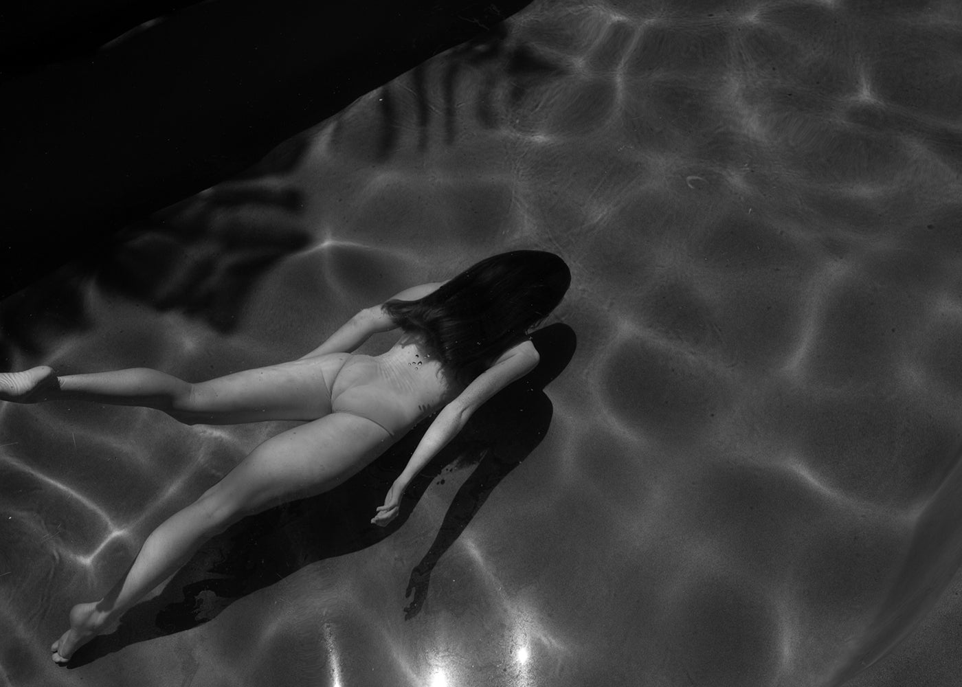 A person lies face down in the shallow water of a swimming pool, wearing nothing, with shadows and light ripples reflecting on the surface. Captured in black and white, Francesca Owen's evocative image titled "Glide" is printed with archival pigments on fine art paper.