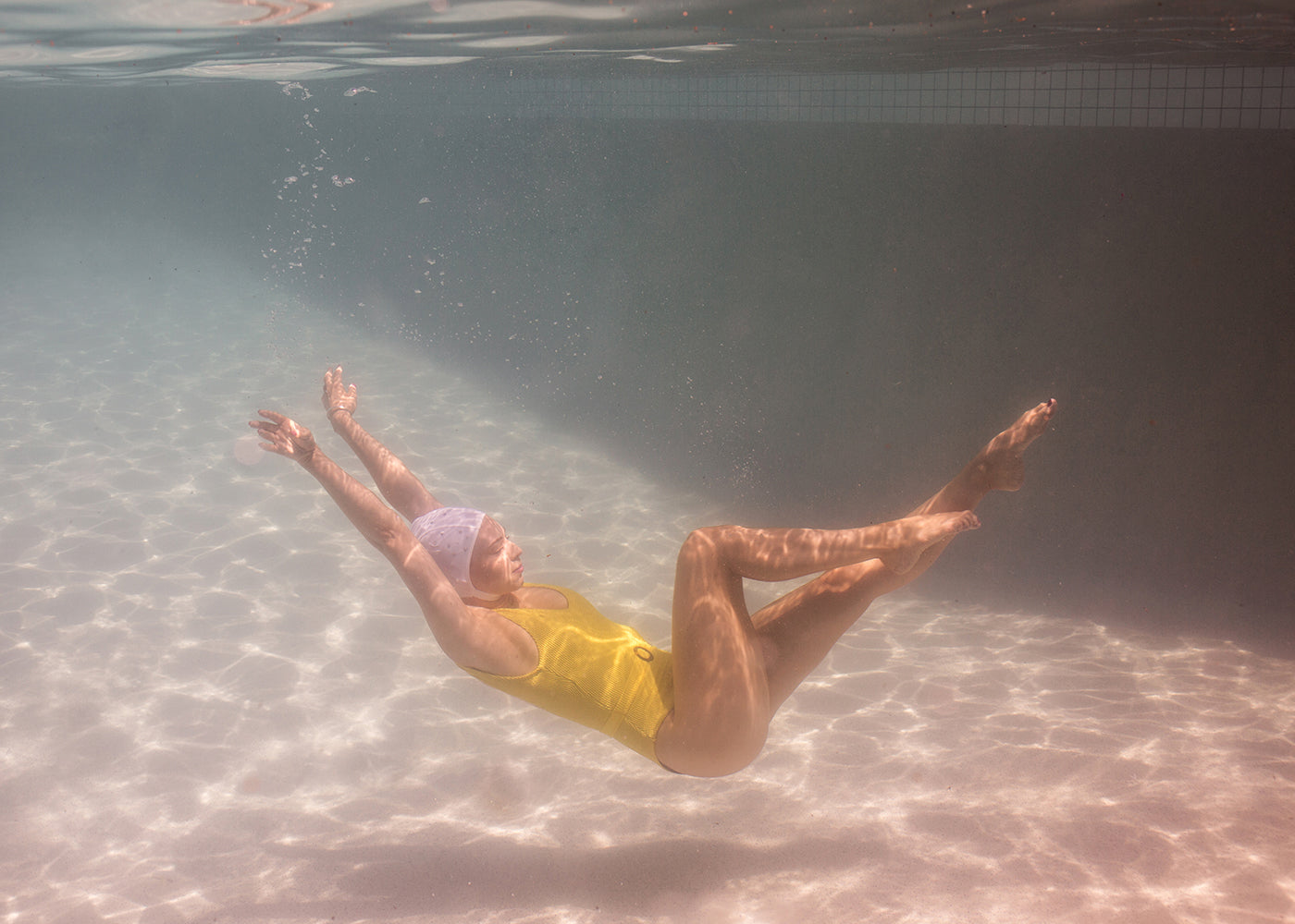 A person wearing a yellow swimsuit and white swimming cap is floating underwater in a swimming pool, with arms outstretched and legs bent. Sunlight filters through the water, creating a rippled effect on the pool floor and walls, resembling Francesca Owen’s "Halcyon Daze Part 1" artwork created with archival pigments on fine art paper.