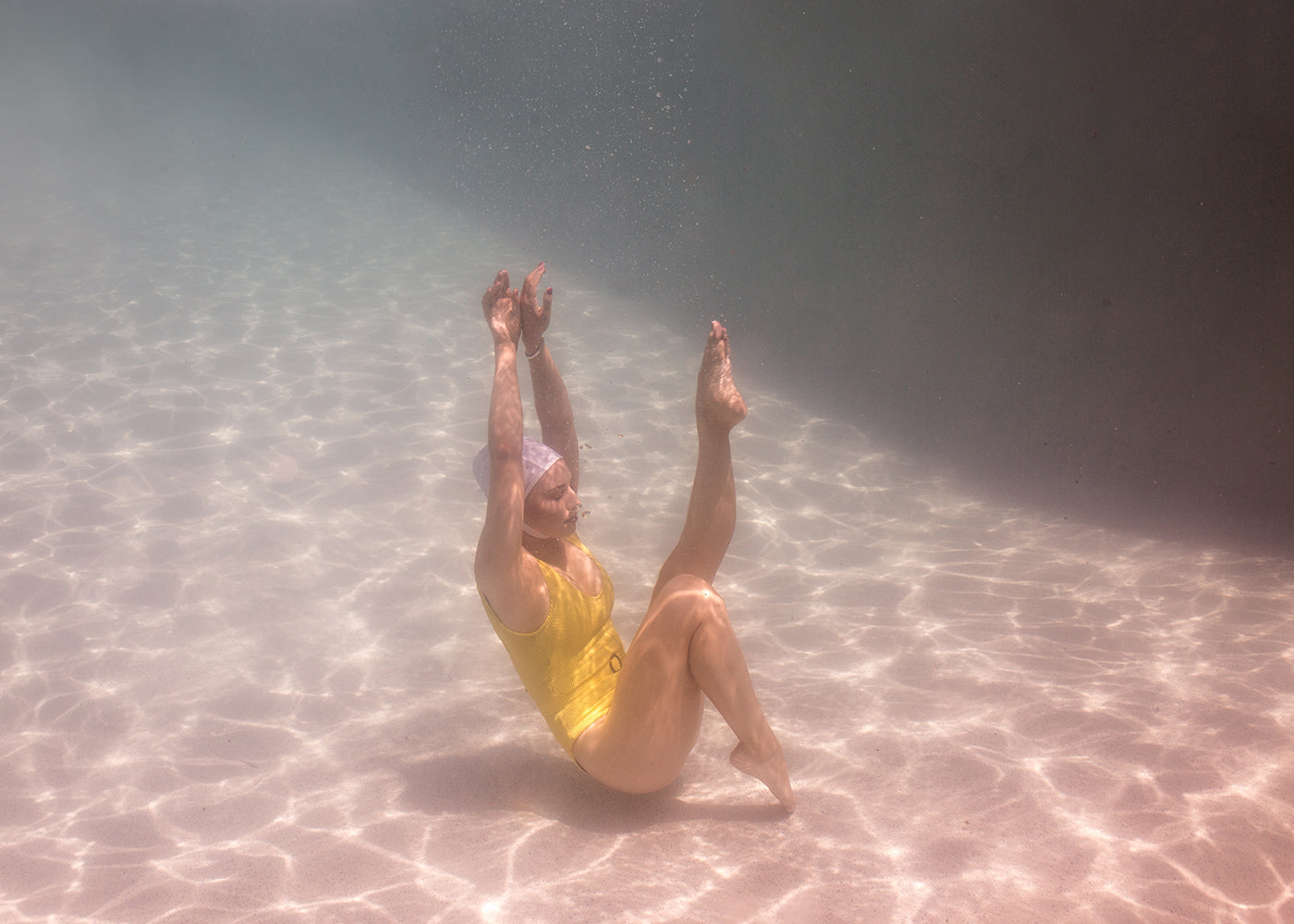 In "Halcyon Daze Part 3" by Francesca Owen, a woman in a yellow swimsuit and swim cap gracefully performs a synchronized swimming pose underwater. Her legs are raised and bent at the knees, while her arms extend upward. Sunlight filters through the water, creating a shimmering effect on the pool floor—perfectly captured as fine art worthy of archival pigments.