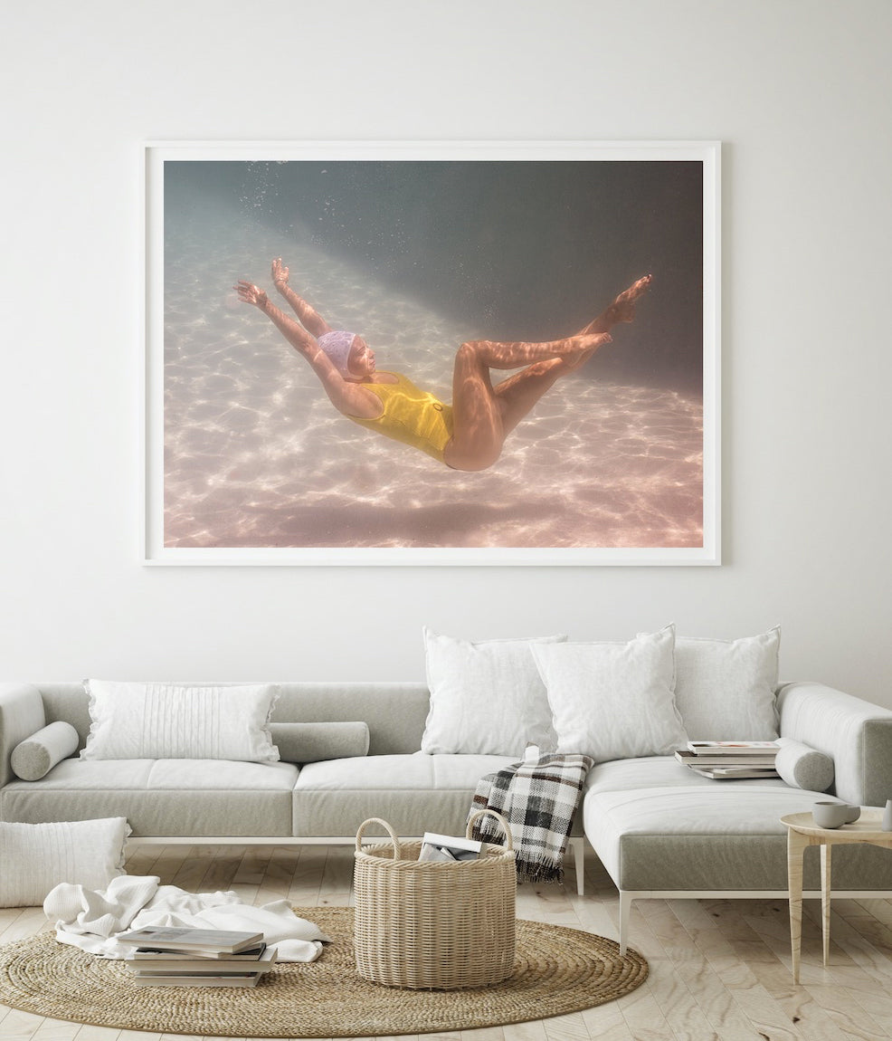 A minimalist living room features a large framed photograph titled "Halcyon Daze Part 1" by Francesca Owen above a modern light gray sofa. The fine art paper photograph, printed with archival pigments, portrays an underwater scene of a person in a yellow swimsuit floating gracefully. The room is decorated with white pillows, a plaid throw, and a woven basket.