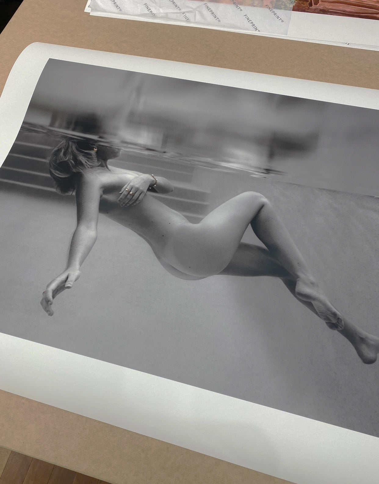 The fine art print titled "Unveiled 05" by Francesca Owen features a black and white photograph of a person swimming underwater in a horizontal position, with one arm extended forward and the other resting on their chest. The surface of the water creates a reflective effect above their body. Presented on a tan surface, this print uses archival pigments for lasting quality.
