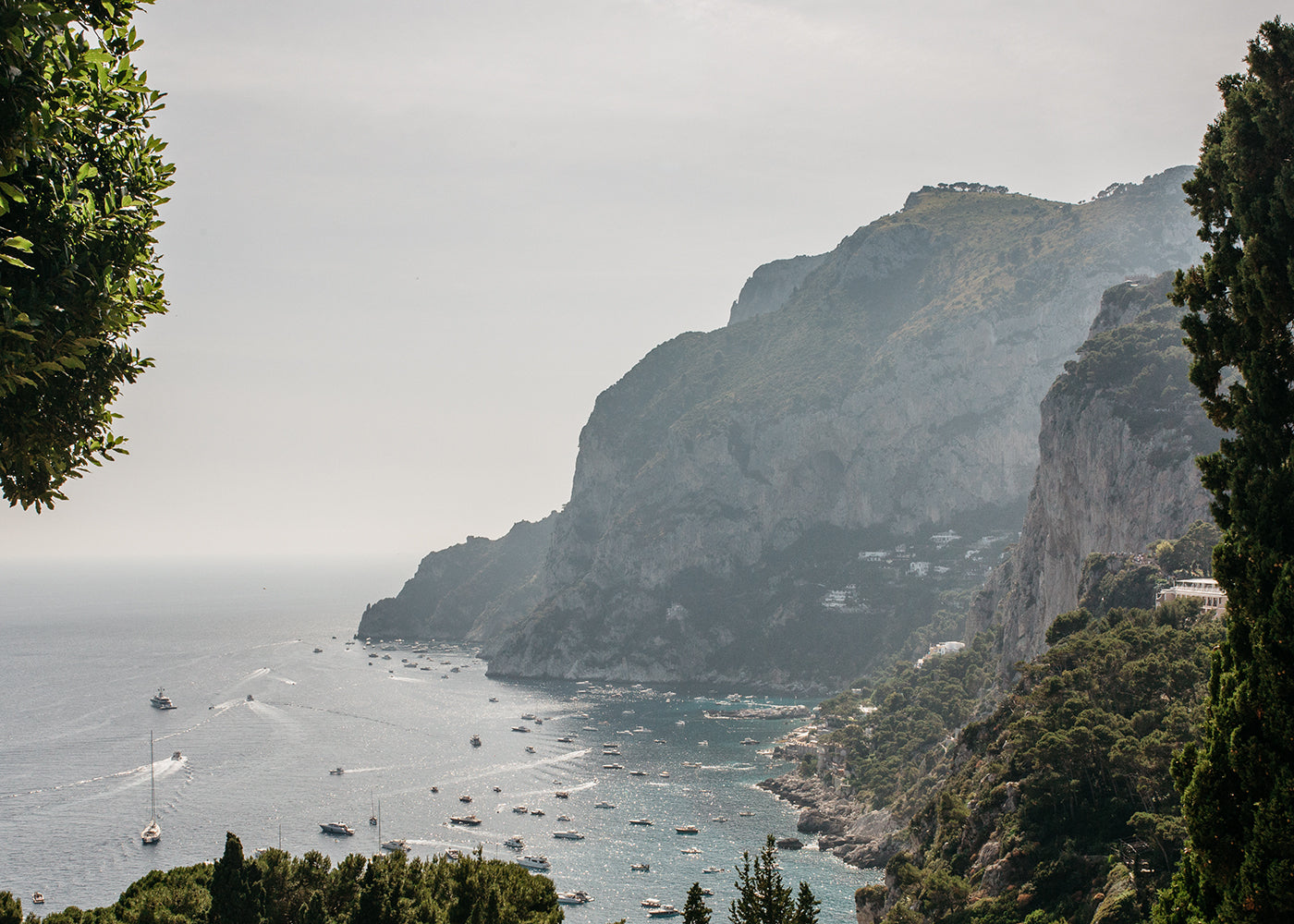 Experience the serene and tranquil scene of 'Capri Rooftops' by Francesca Owen, capturing a vast expanse of sea dotted with numerous boats under a scenic coastal view. Rugged cliffs rise majestically from the shoreline, partially covered with dense greenery. This breathtaking artwork is rendered in stunning detail using archival pigments for fine art prints, each accompanied by a certificate of authenticity.