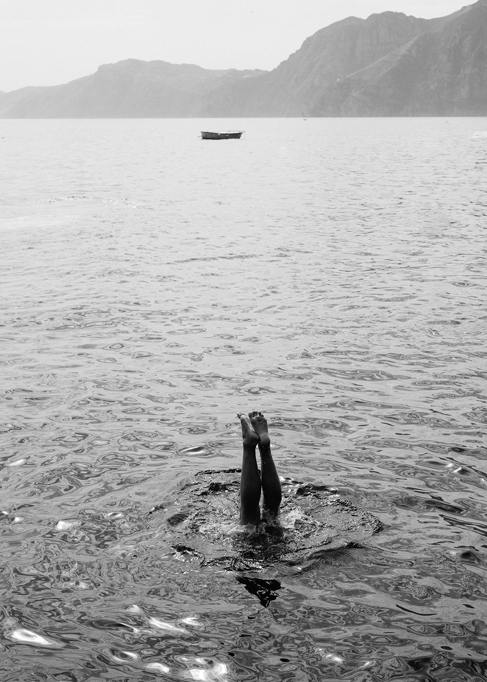 Capri Tippy Toes" by Francesca Owen captures a striking black and white image of a person performing a handstand in the water, with only their legs and feet visible above the surface. In the background, distant mountains and a solitary boat float on calm waters. This piece is available in custom sizes with archival pigments and includes a certificate of authenticity.