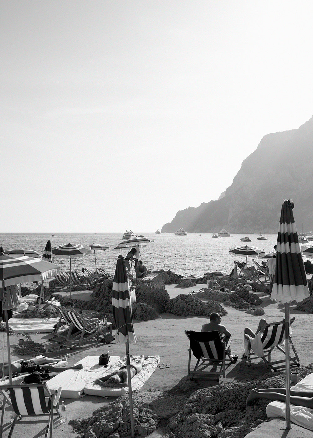 La Fontelina #2 by Francesca Owen captures a black and white beach scene where people lounge on chairs beneath striped umbrellas. The picturesque beach, with its rocky outcrops, is set against a backdrop of the sea, featuring boats anchored in the distance and a mountainside on the right. This exquisite piece is printed on fine art paper using archival pigments and is available for worldwide shipping.