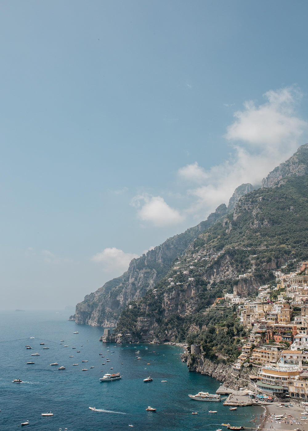 The artwork, "Positano #1" by Francesca Owen, vividly depicts a cliffside town where vibrant buildings are stacked atop one another, overlooking a clear blue sea speckled with numerous boats. The scene features a clear sky with a few wispy clouds and green hills rising behind the town. This piece is available in custom sizes and is printed with archival pigments to ensure lasting beauty.