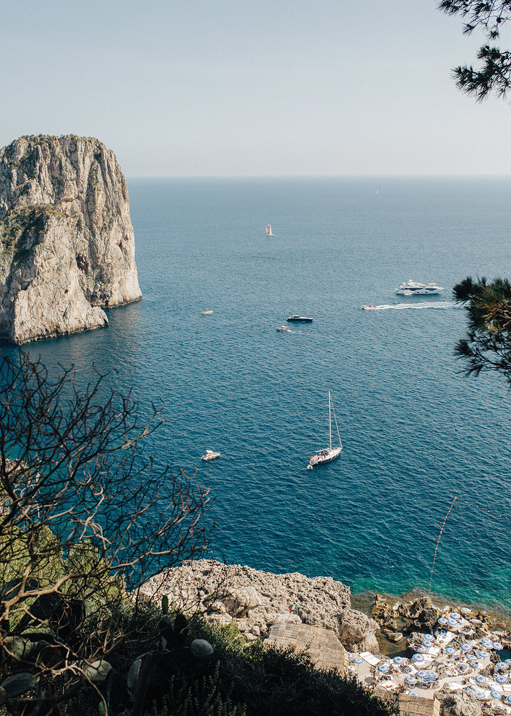 Capture the scenic view of the coastline in "Sailing Capri #4" by Francesca Owen, where a large cliff juts into the calm blue sea. Several boats, including a sailboat and yachts, glide on the water. Rocky shores and lush greenery are in the foreground under a clear, sunny sky—ideal for creating fine art prints with archival pigments for worldwide shipping.