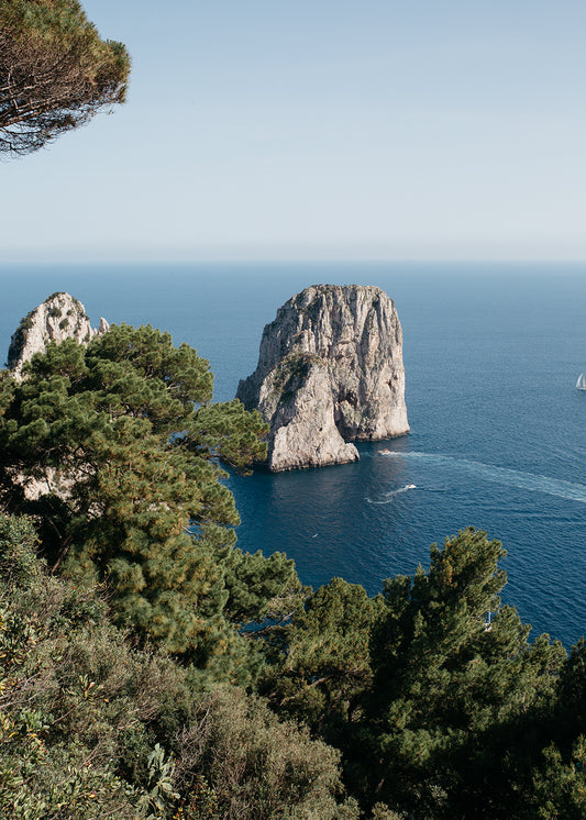Behold the breathtaking "Sailing Capri #1" by Francesca Owen: a scenic view of two majestic rock formations emerging from the azure sea, ringed by verdant green trees in the foreground. The clear, brilliant sky enhances the coastal landscape's natural allure—masterfully captured using archival pigments on Fine Art paper, available in custom sizes.