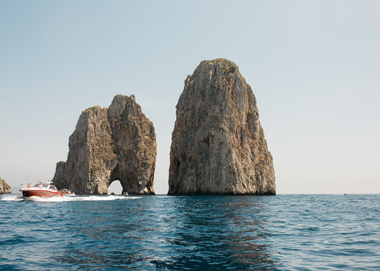 In "Sailing Capri #2" by Francesca Owen, a vibrant red and white speedboat races past the iconic Faraglioni rock formations in Capri, Italy, beneath a clear blue sky. The sea is tranquil, and a small arch can be seen in one of the towering rocks, captured with precision as if printed with archival pigments on fine art paper.