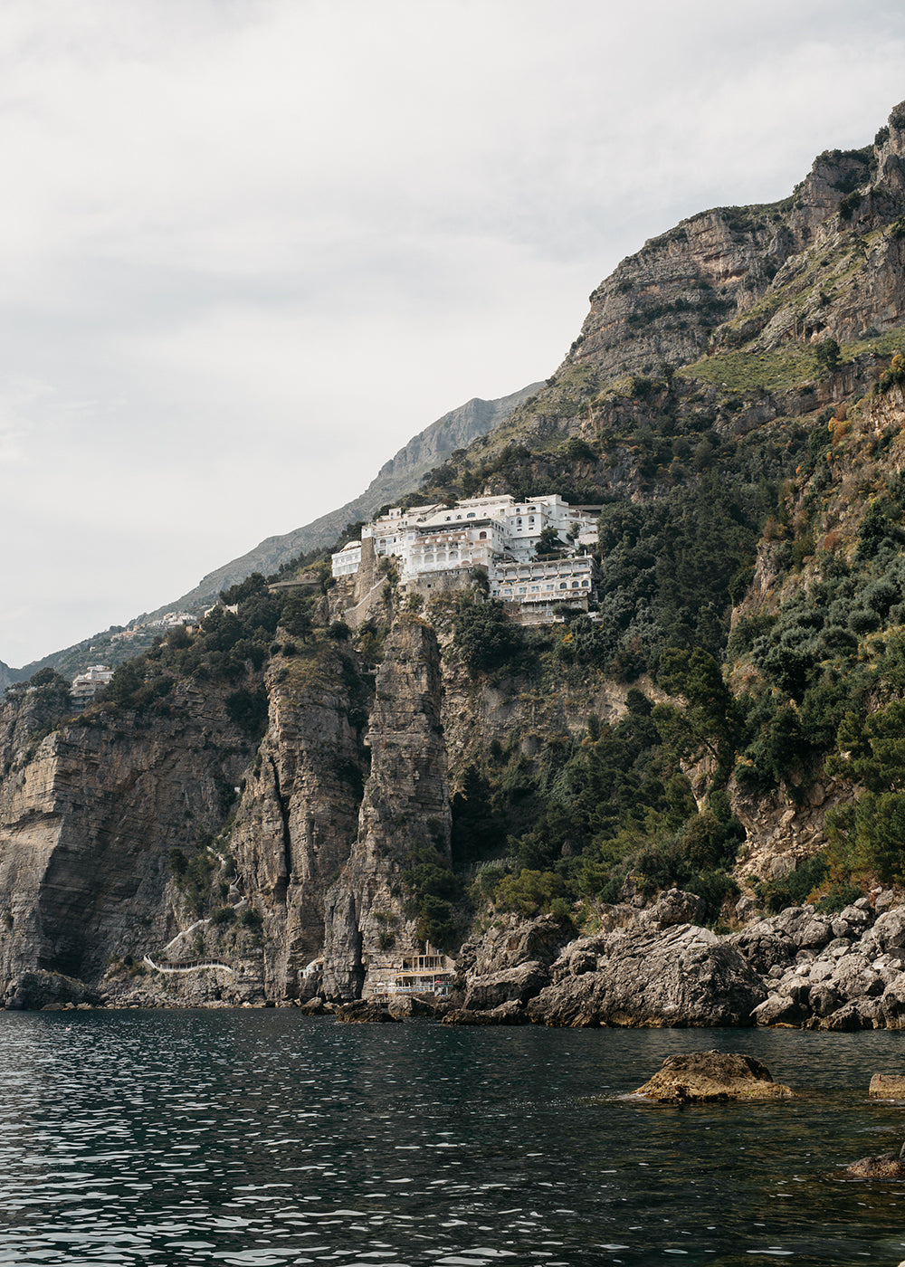Experience the mesmerizing view of a coastal cliff featuring rugged terrain and lush greenery, beautifully captured on fine art paper with archival pigments, in "Seaside in Praiano" by Francesca Owen. Several white buildings are perched near the cliff's summit, overlooking the serene blue sea below. The mostly cloudy sky adds a touch of drama to this masterpiece, which is available for worldwide shipping.
