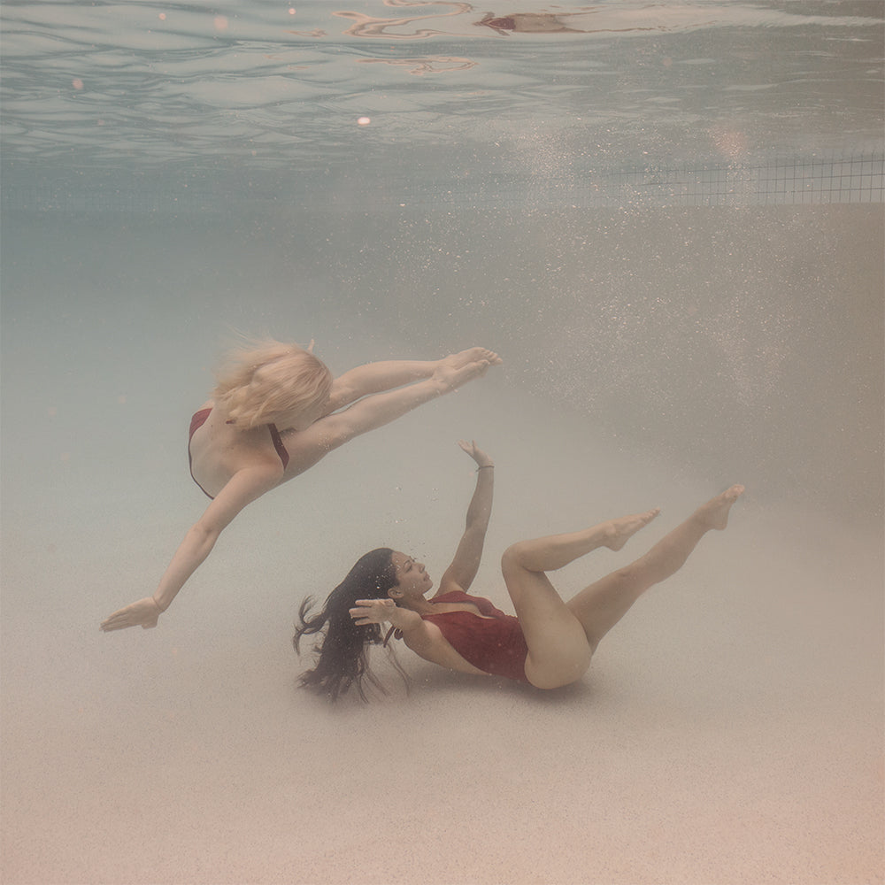 In the fine art print "Opposites Attract #2" by Francesca Owen, two women in red swimsuits are performing an underwater dance routine. One is gracefully flipping mid-air with outstretched arms and legs, while the other appears to be floating upwards with her hair splayed out. Bubbles and soft lighting surround them, creating a mesmerizing visual experience capture beautifully in this artwork.