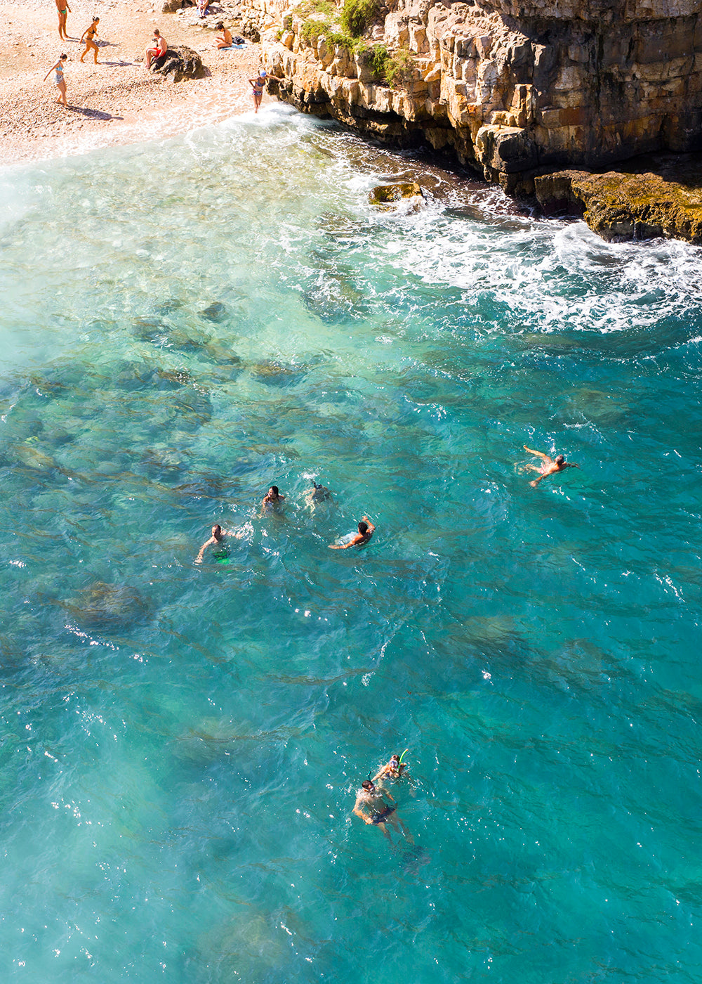 An aerial view of a coastal area with people swimming in clear, turquoise water. The rocky shoreline with some greenery is visible at the top, and a sandy beach occupies the upper left corner where a few people sunbathe and stand. Waves crash gently against the rocks, ideal for capturing in fine art prints using archival pigments. This stunning scene is perfectly encapsulated in "A Polignano a Mare" by Francesca Owen.