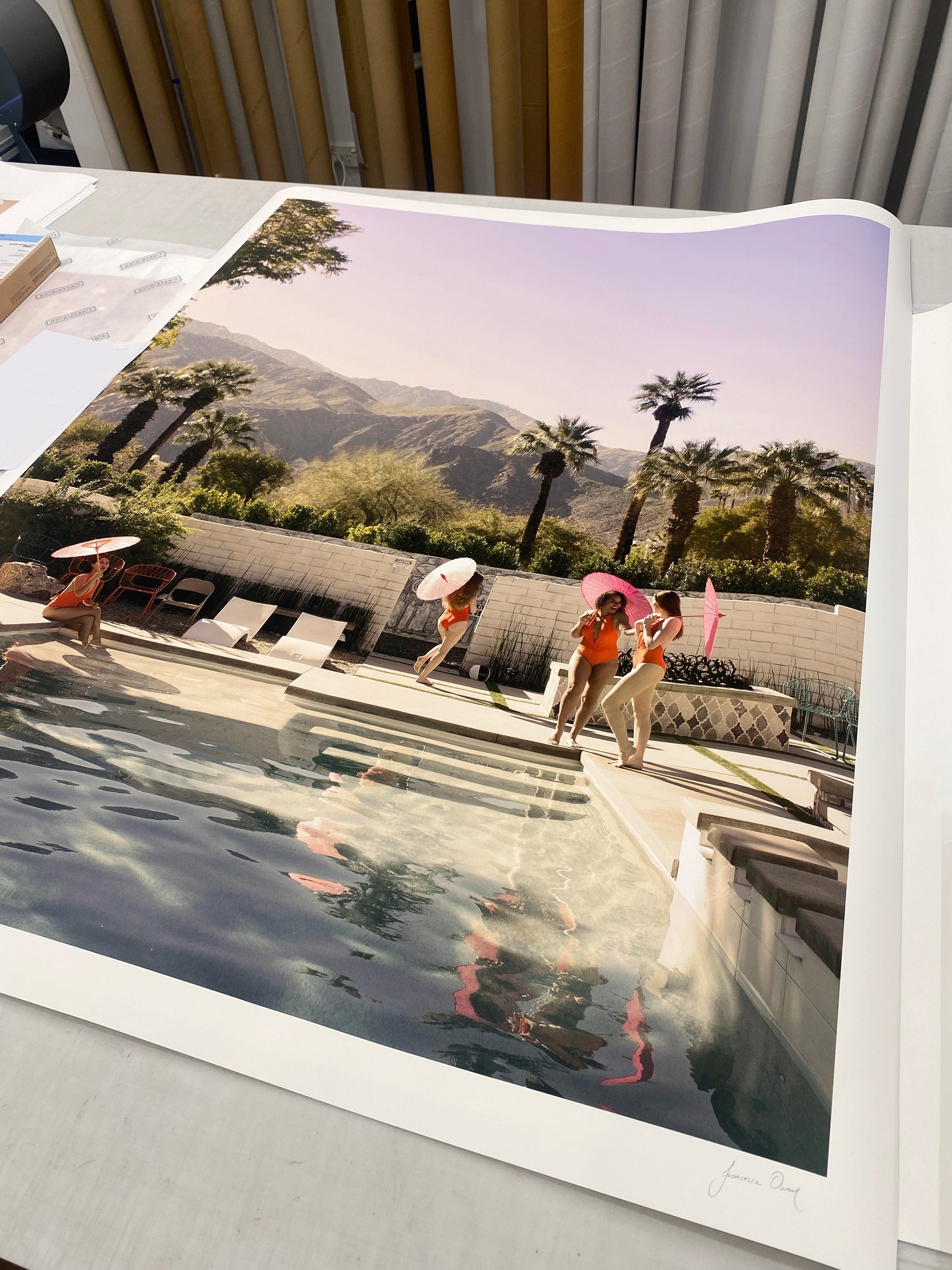 A retro-style photograph titled "Poolside Mermaids" by Francesca Owen showcases three women in orange swimsuits standing by a pool, each holding a pink parasol. The scene is beautifully set against a backdrop of palm trees, a white wall, and distant mountains under a clear sky. This vibrant image is available as limited edition prints on fine art paper.