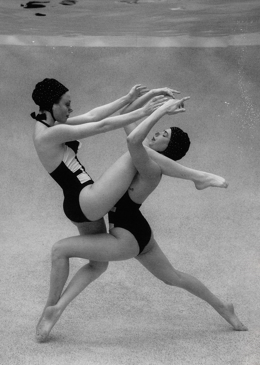 Black and white image of two synchronized swimmers underwater. Both are dressed in swimsuits and swim caps, gracefully performing an intricate, acrobatic routine that involves extended legs and outstretched arms, showcasing their balance and coordination. Titled 'Palm Springs Duo #1,' this captivating photograph by Francesca Owen is printed on fine art paper with archival pigments.