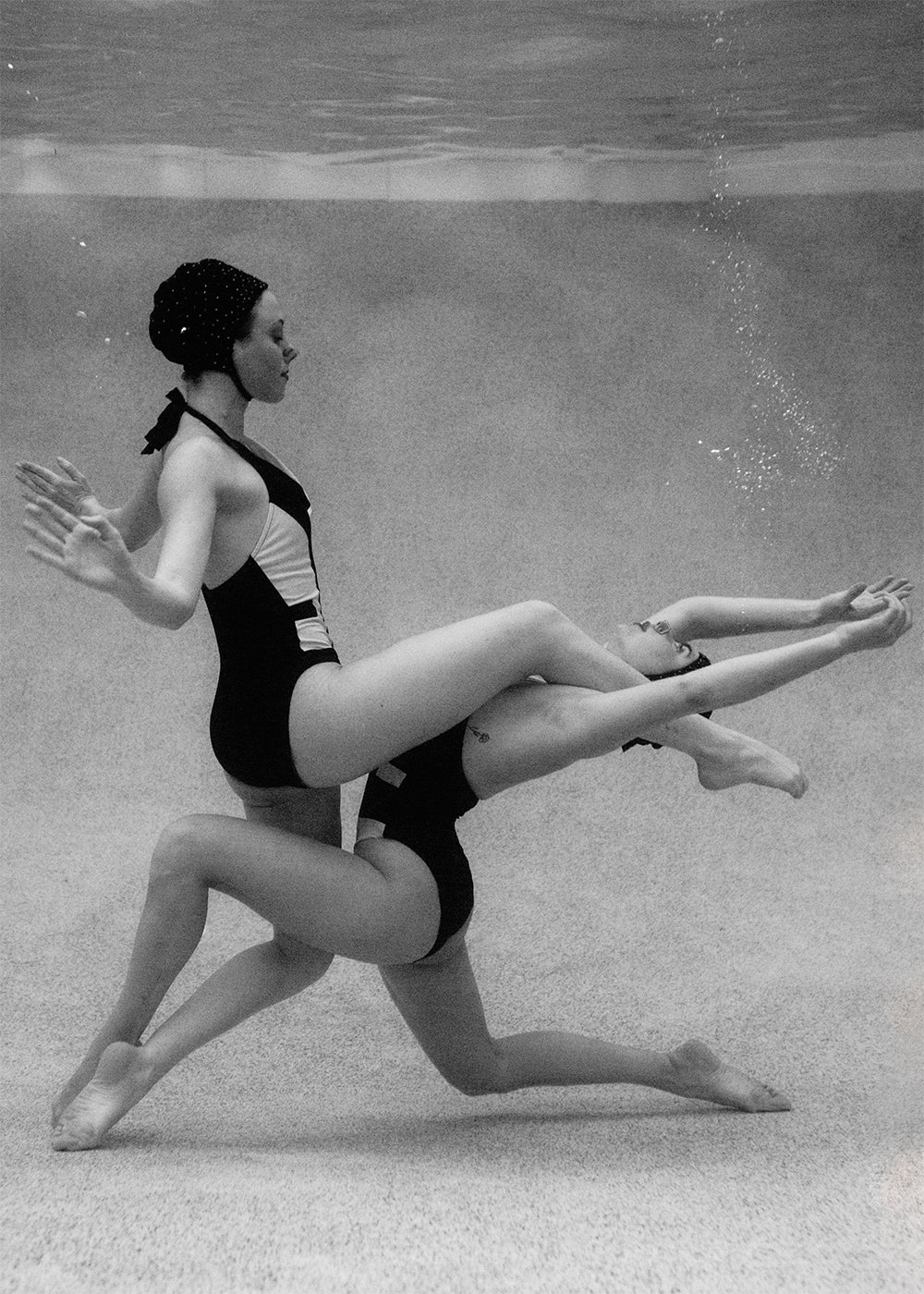 Two synchronized swimmers perform an underwater routine in black and white. One balances on the other's raised knee, extending her arms and legs gracefully. Both wear matching swimsuits and swim caps as air bubbles rise from their movements. Perfect for custom framing, this scene, titled "Palm Springs Duo #2" by Francesca Owen, is a timeless art print.
