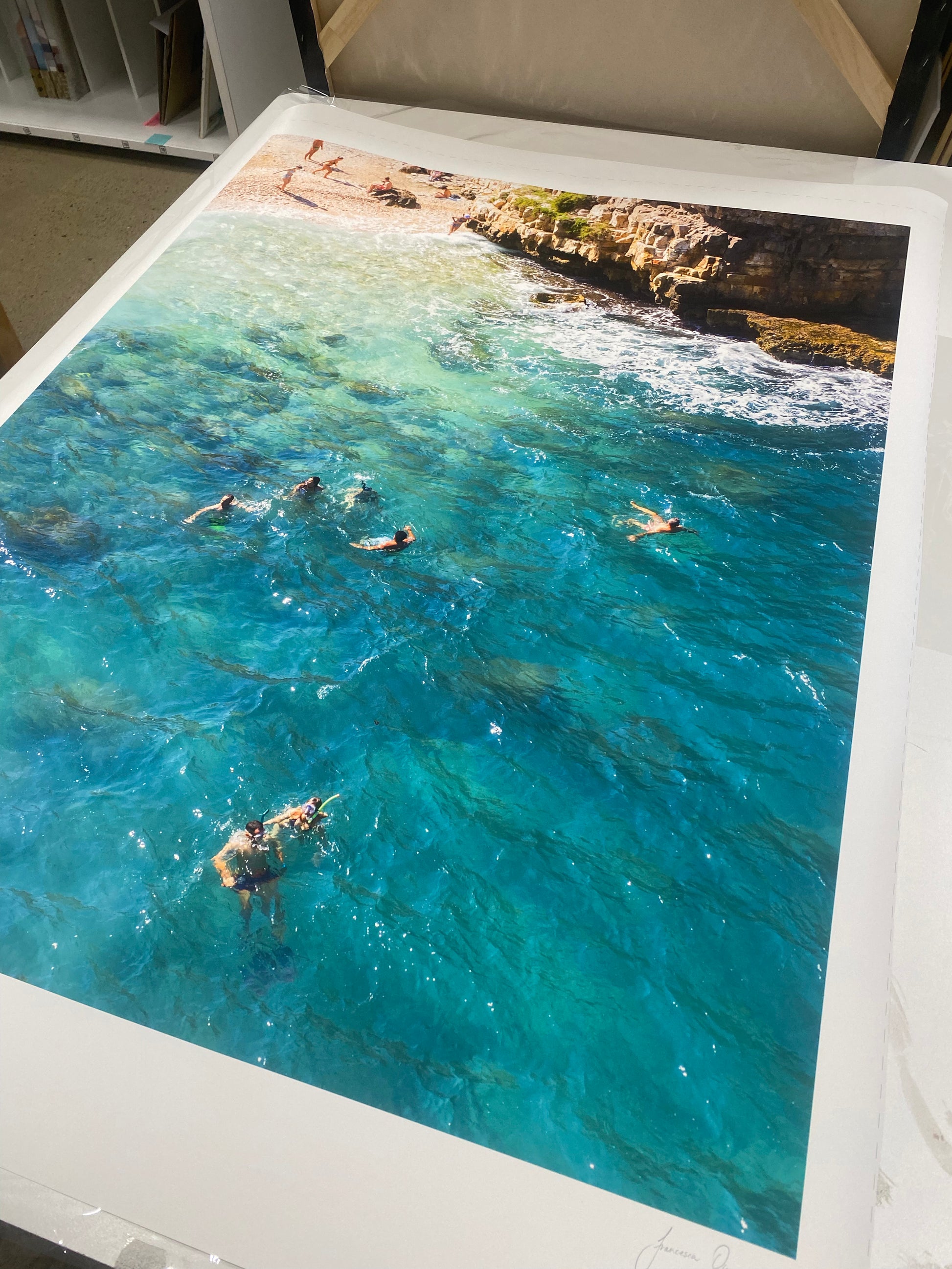 A Polignano a Mare, a framed photograph by Francesca Owen, produced with archival pigments, hangs on a quilted wall. The picture depicts multiple people swimming in the clear blue waters of a rocky beach. The scene is vibrant with shades of blue and green, capturing a sunny day at the coast.