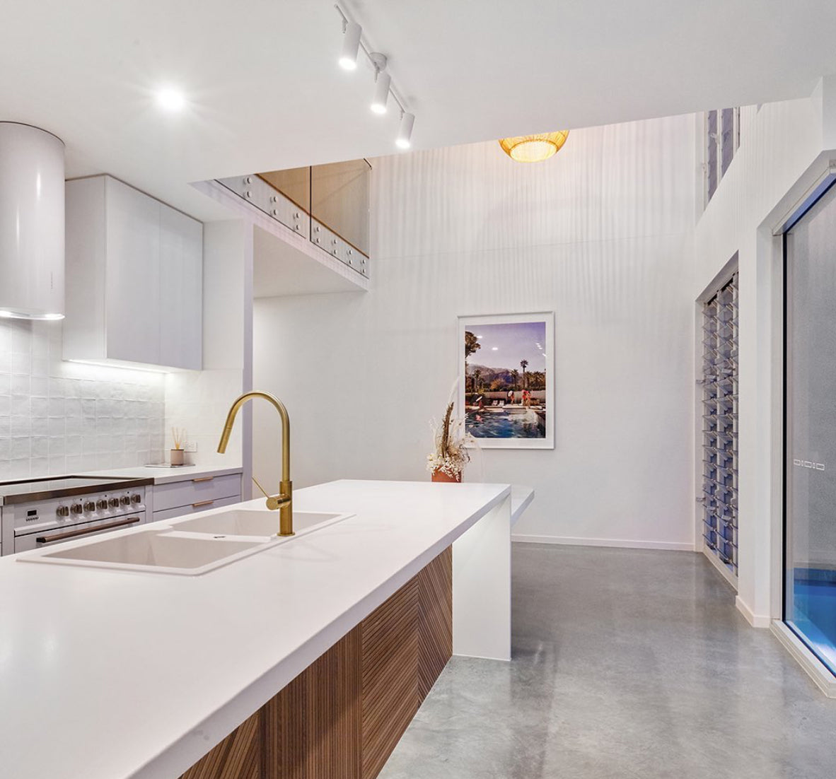 Modern kitchen with a white island featuring a sleek gold faucet and integrated sink. The space is illuminated by both built-in lighting and natural light from high windows. Minimalist decor includes "Poolside Mermaids," a hand-signed, framed picture by Francesca Owen with a certificate of authenticity on a white wall, along with wooden accents.