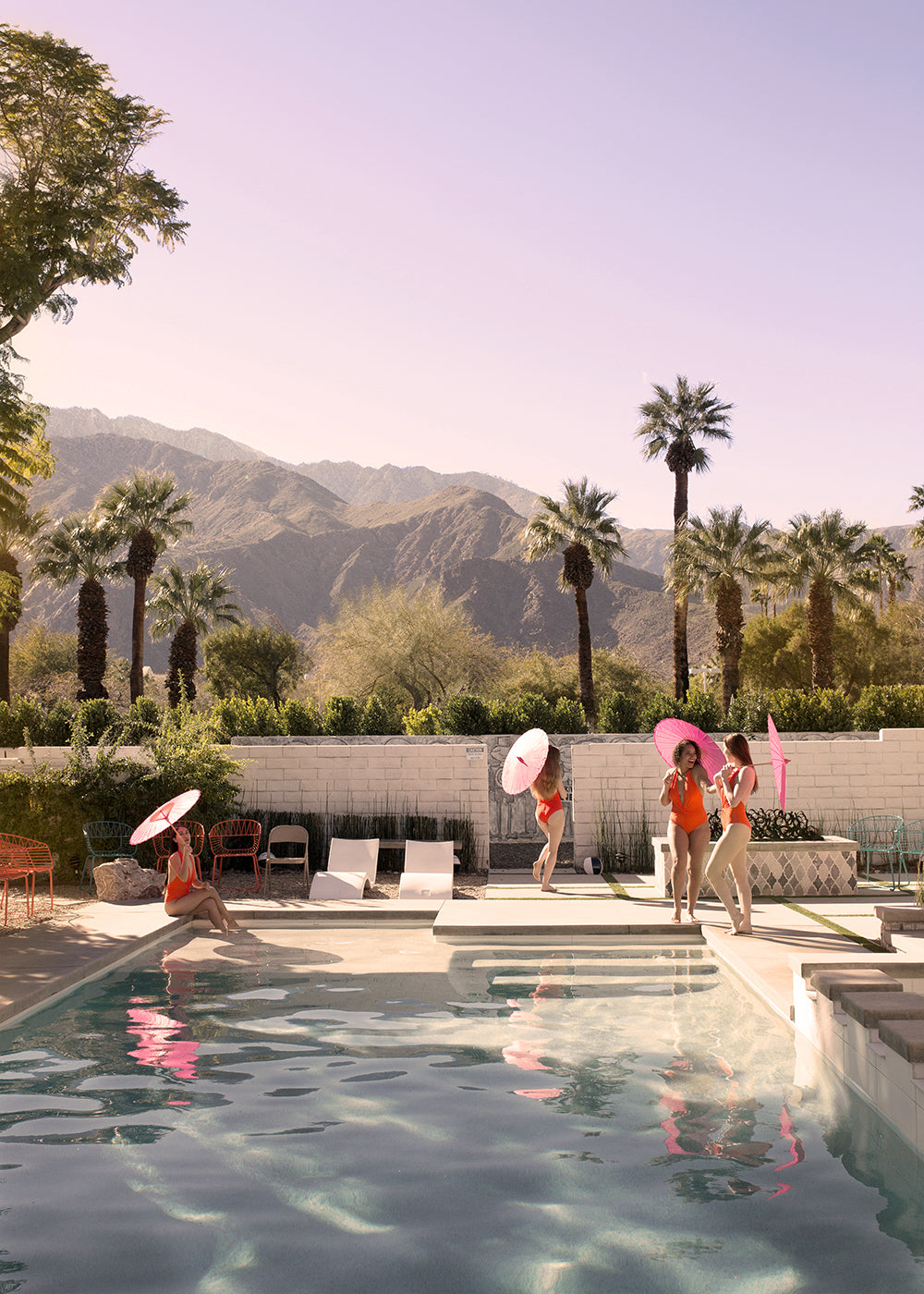 A serene poolside scene from "Poolside Mermaids" by Francesca Owen depicts several individuals, each holding hand-signed pink parasols, relaxing and engaging in conversation. The setting is surrounded by lush palm trees and a mountainous backdrop under a clear sky. Some people are seated while others stand near the pool.