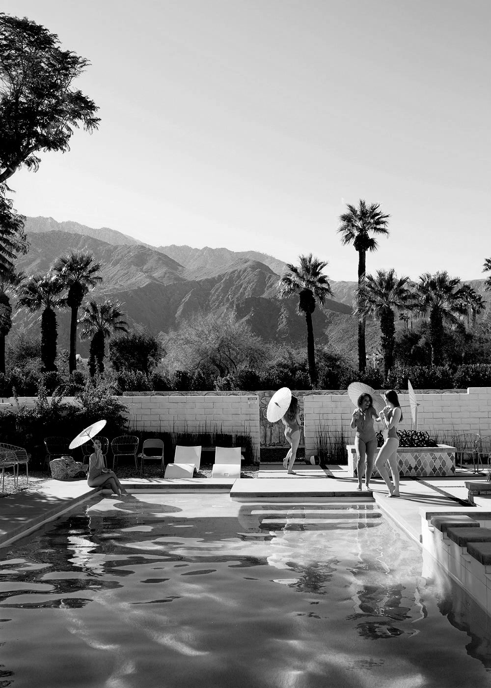 The limited edition print "Poolside Mermaids B&W" by Francesca Owen beautifully captures four people by a pool in a picturesque desert setting with mountain views and palm trees. Two individuals hold parasols, while others lounge on pool chairs. Created using archival pigments, the photo exudes a relaxed and sunny atmosphere.
