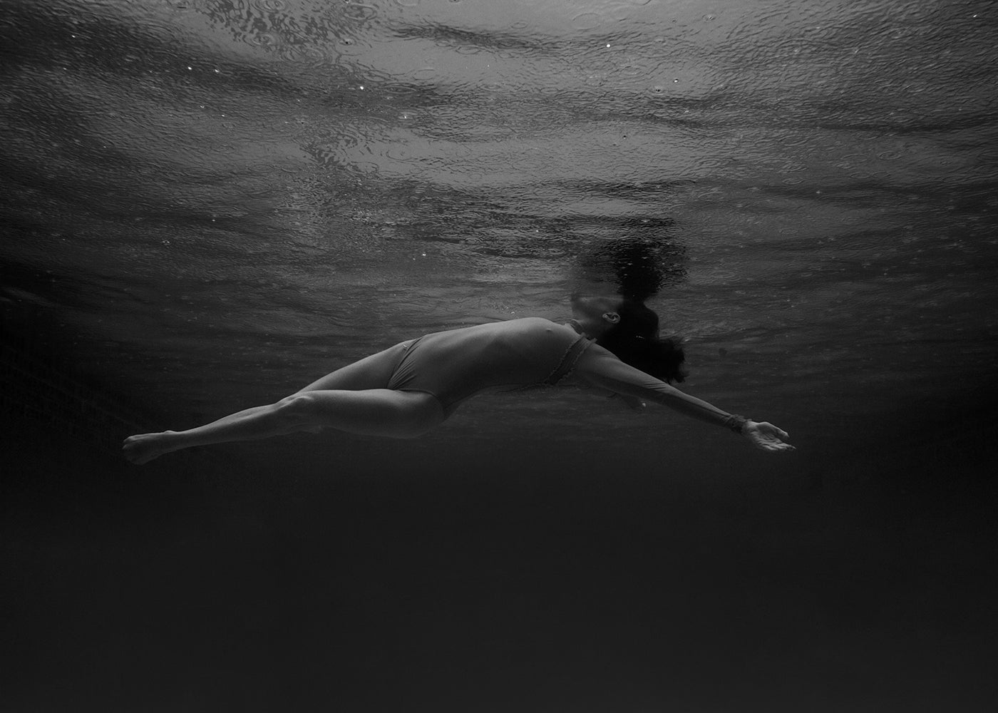 In the black and white artwork titled "Red Head Series #10" by Francesca Owen, a person in a bikini is captured swimming underwater in a horizontal position with their arms extended backward. The textured effect created by the water's surface above enhances the serene and calm appearance of the subject, beautifully rendered on fine art paper.