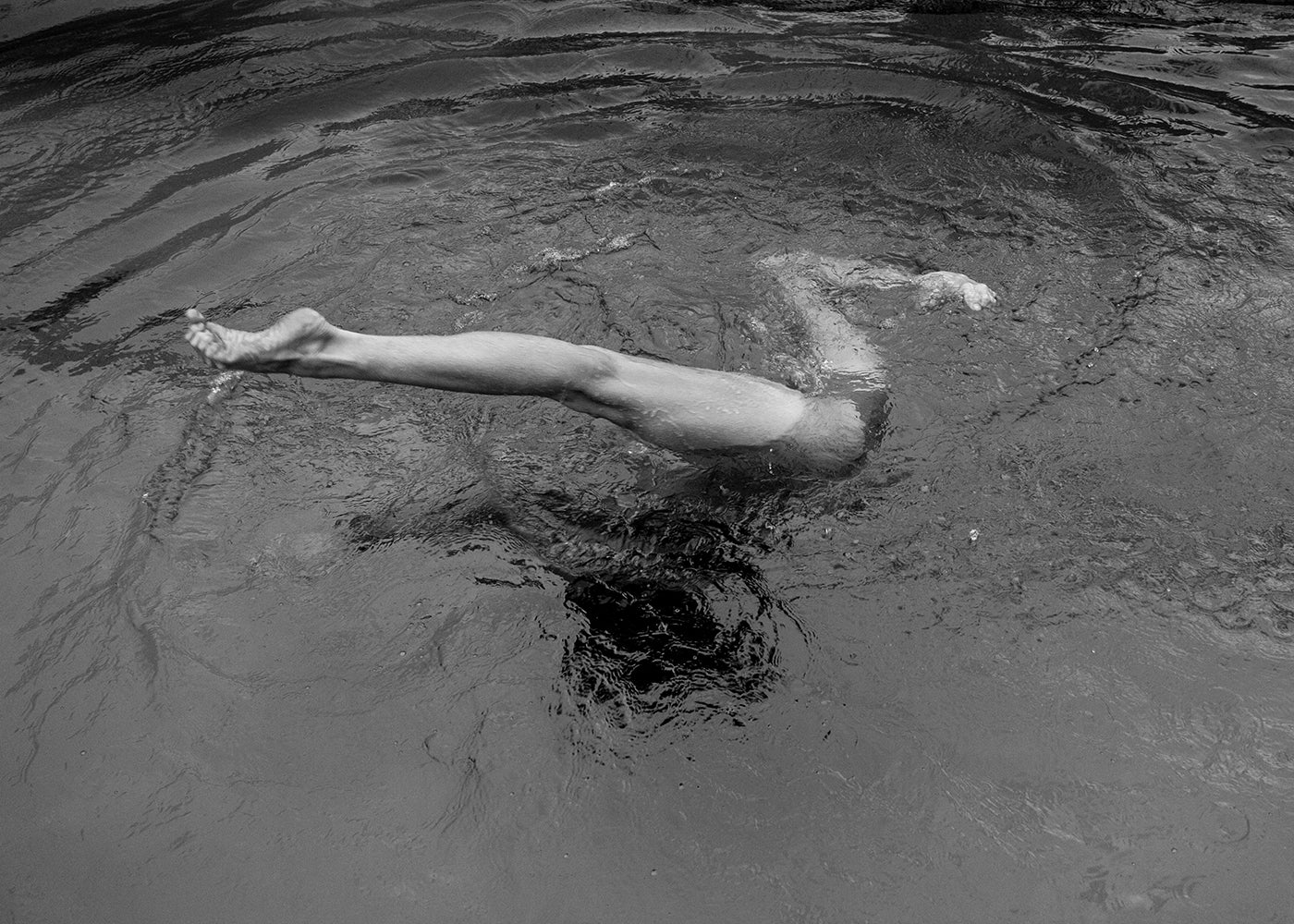 A person is swimming underwater with their legs and feet visible above the water's surface. The black and white image, part of Francesca Owen's Red Head Series #7, captures the ripples around the swimmer using archival pigments that ensure lasting quality, and is available as fine art prints.