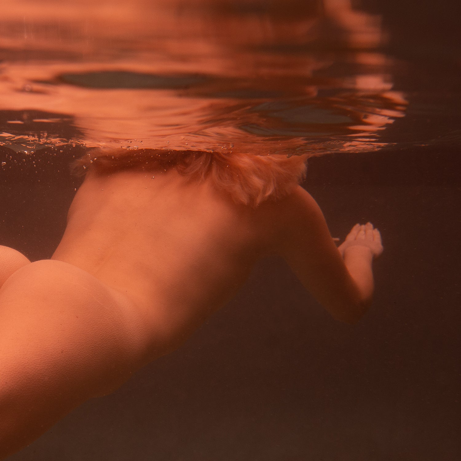 In "Reflect no.3" by Francesca Owen, a person with blonde hair is captured swimming underwater, facing away from the camera. The upper part of their body is submerged while the water above reflects their silhouette. The lighting casts a warm, reddish tint over the scene. This evocative moment is printed on fine art paper using archival pigments and comes with a certificate of authenticity.