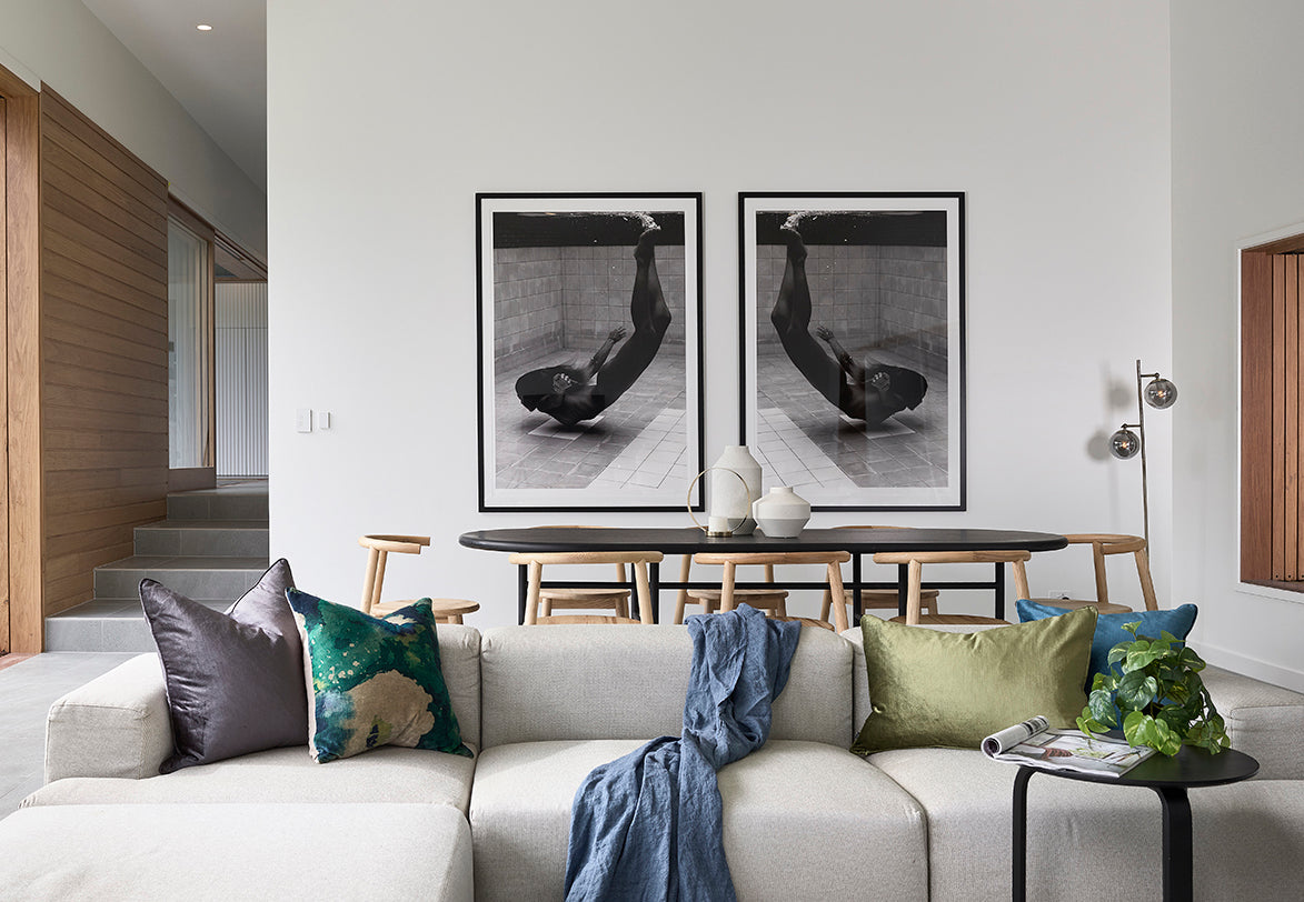 A modern living room boasts the "Fall" sofa from Francesca Owen, adorned with colorful cushions. Behind this light-colored sofa stands a round dining table paired with black chairs. The wall is decorated with two identical framed art prints on fine art paper, adding a touch of elegance. On the left side, a staircase is partially visible.
