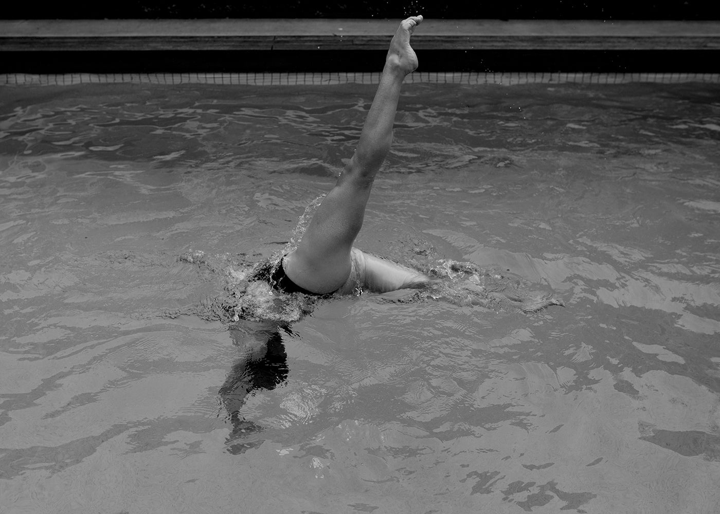 Self Reflection 19 by Francesca Owen features a black and white photo capturing a person performing a leg raise in a swimming pool. The image shows only one leg and part of the body above the water's surface, with the rest submerged. Ripples from the movement are visible in the water, printed on fine art paper using archival pigments for lasting quality.