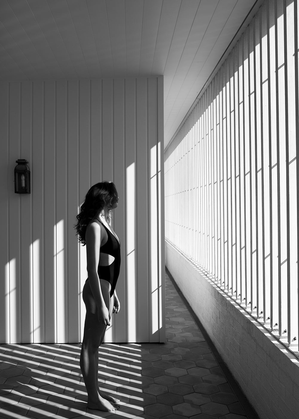 A woman in a swimsuit stands in profile facing the sunlight streaming through vertical bars onto a tiled floor. The strong shadows create striking patterns on the wall and floor, emphasizing the geometric lines of the space. This black-and-white scene, titled "Self Reflection 10" by Francesca Owen, is printed on fine art paper with archival pigments and comes with a certificate of authenticity.