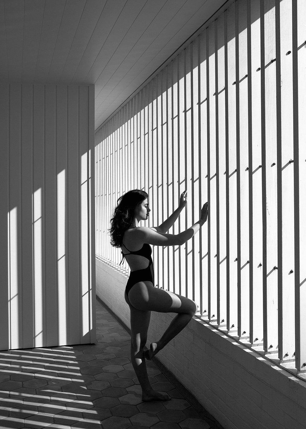 A woman in a black swimsuit stands under sunlight streaming through vertical bars casting shadows. She has one leg raised and hands positioned against the bars. The background is composed of white walls and a patterned floor, all expertly captured by Francesca Owen’s "Self Reflection 09" on fine art paper with archival pigments, ensuring timeless elegance.