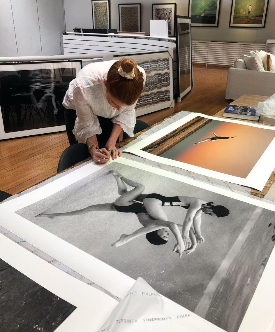 A person with red hair tied in a bun leans over a table, signing large photographic prints in a gallery. The prints, created using archival pigments on fine art paper, include various images such as "Palm Springs Duo #1" by Francesca Owen, featuring monochrome ballet dancers and vibrant scenes of someone diving into water. Art frames and prints adorn the walls.