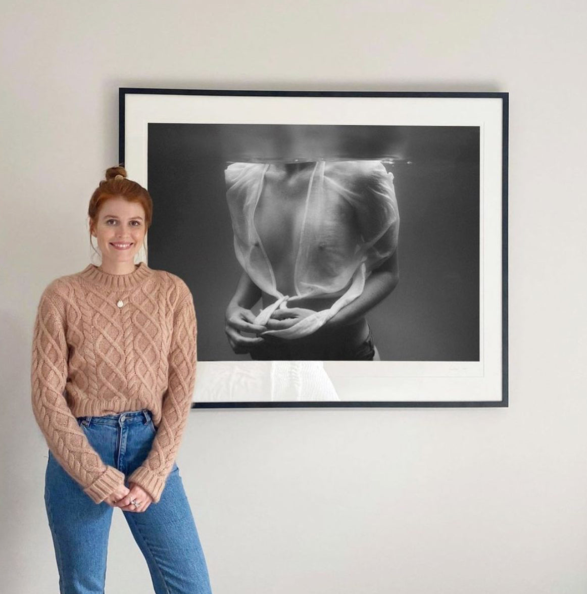 A woman with red hair is standing and smiling in front of "Softness #2," a wall-mounted black-and-white photograph by Francesca Owen, printed on fine art paper and depicting a partially submerged person covered by translucent fabric. She is wearing a tan cable-knit sweater and blue jeans.