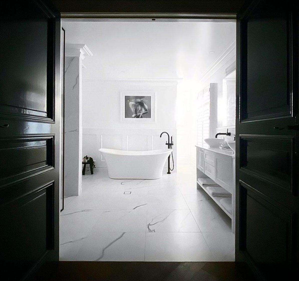 A framed black and white fine art print from Francesca Owen, titled "Softness #1 B&W," hangs on a white wall. It depicts a partially submerged person in water. Next to the photo is a window with a view of a parked car and a brick house across the street. A lacy white garment is seen hanging on the left side of the image, enhanced by archival pigments for lasting beauty.