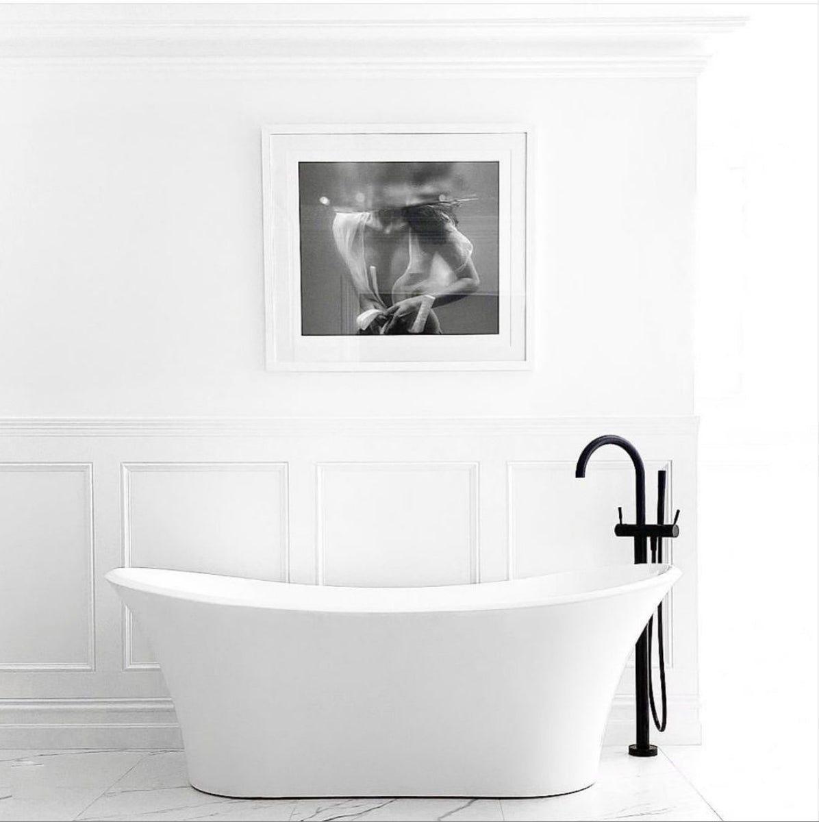 A framed black and white fine art print from Francesca Owen, titled "Softness #1 B&W," hangs on a white wall. It depicts a partially submerged person in water. Next to the photo is a window with a view of a parked car and a brick house across the street. A lacy white garment is seen hanging on the left side of the image, enhanced by archival pigments for lasting beauty.