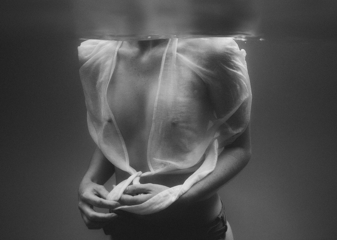 Softness #2 by Francesca Owen is a black and white underwater photograph of a person with a sheer, translucent top, partially submerged, covering their face and head. Their hands grip the hem of the top at their midsection. Printed on fine art paper using archival pigments, this piece exudes an ethereal and mysterious vibe.