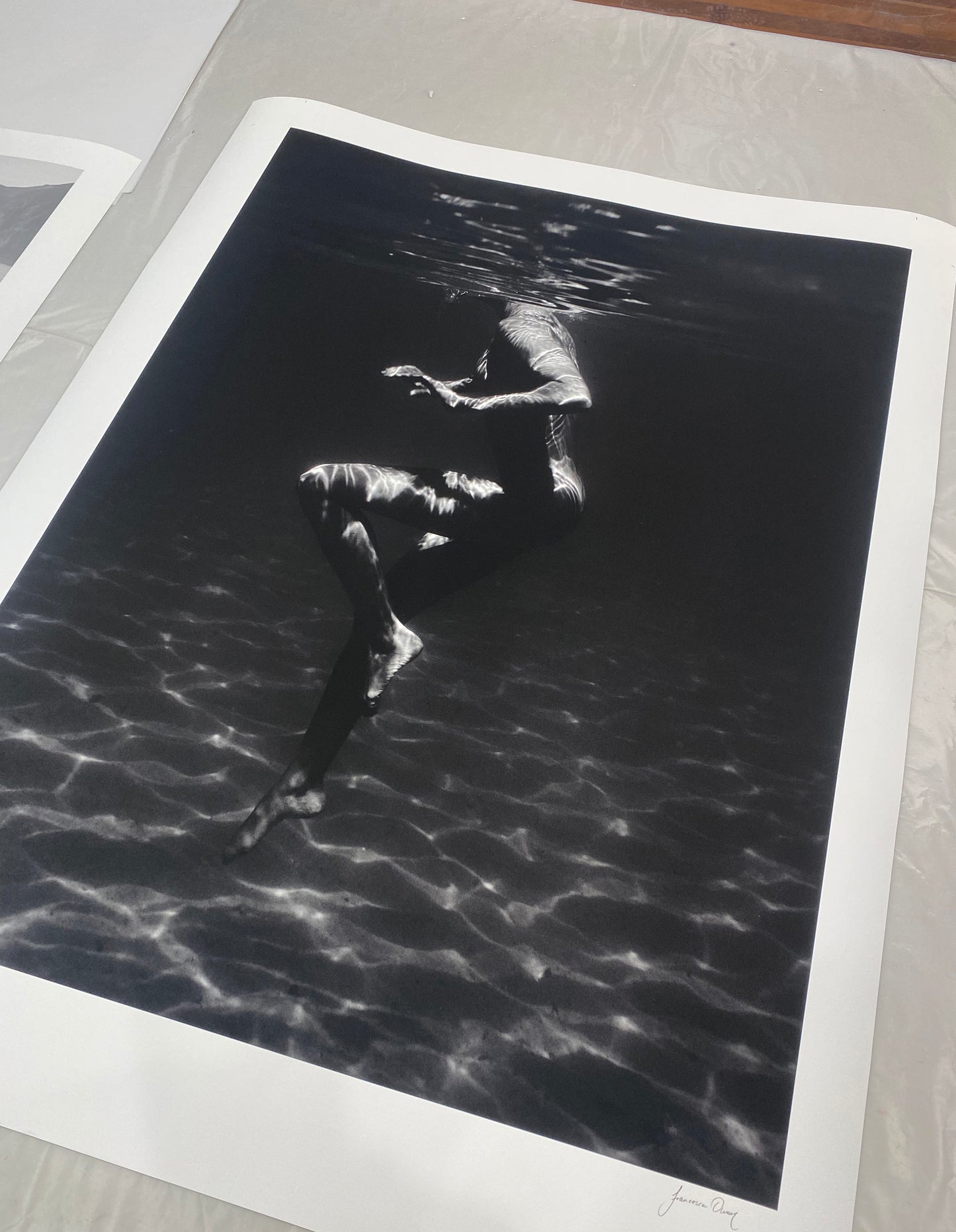"Still Waters #1" by Francesca Owen showcases a mesmerizing photograph of a person submerged in water with light reflections creating intricate patterns on their body. The image captures the motion and fluidity of being underwater and is printed on fine art paper using archival pigments. Available for worldwide shipping.