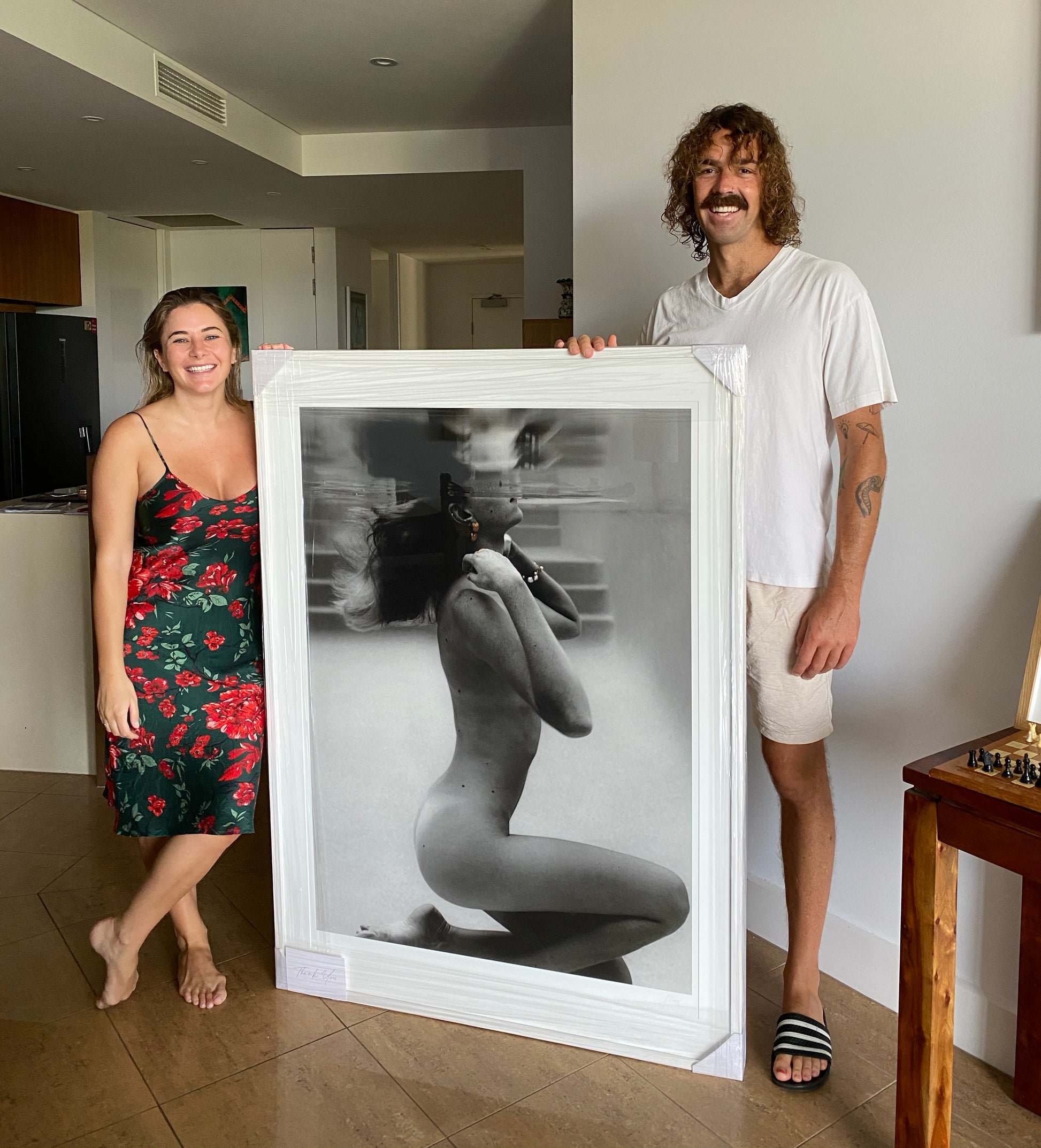 An artful black-and-white photograph from the "UNVEILED" collection by Francesca Owen, printed on fine art paper with archival pigments, graces a white shiplap wall. The image captures a partially clothed person in a seated partial profile. Below this framed Limited Edition print, a sofa adorned with grey and white cushions is partially visible.