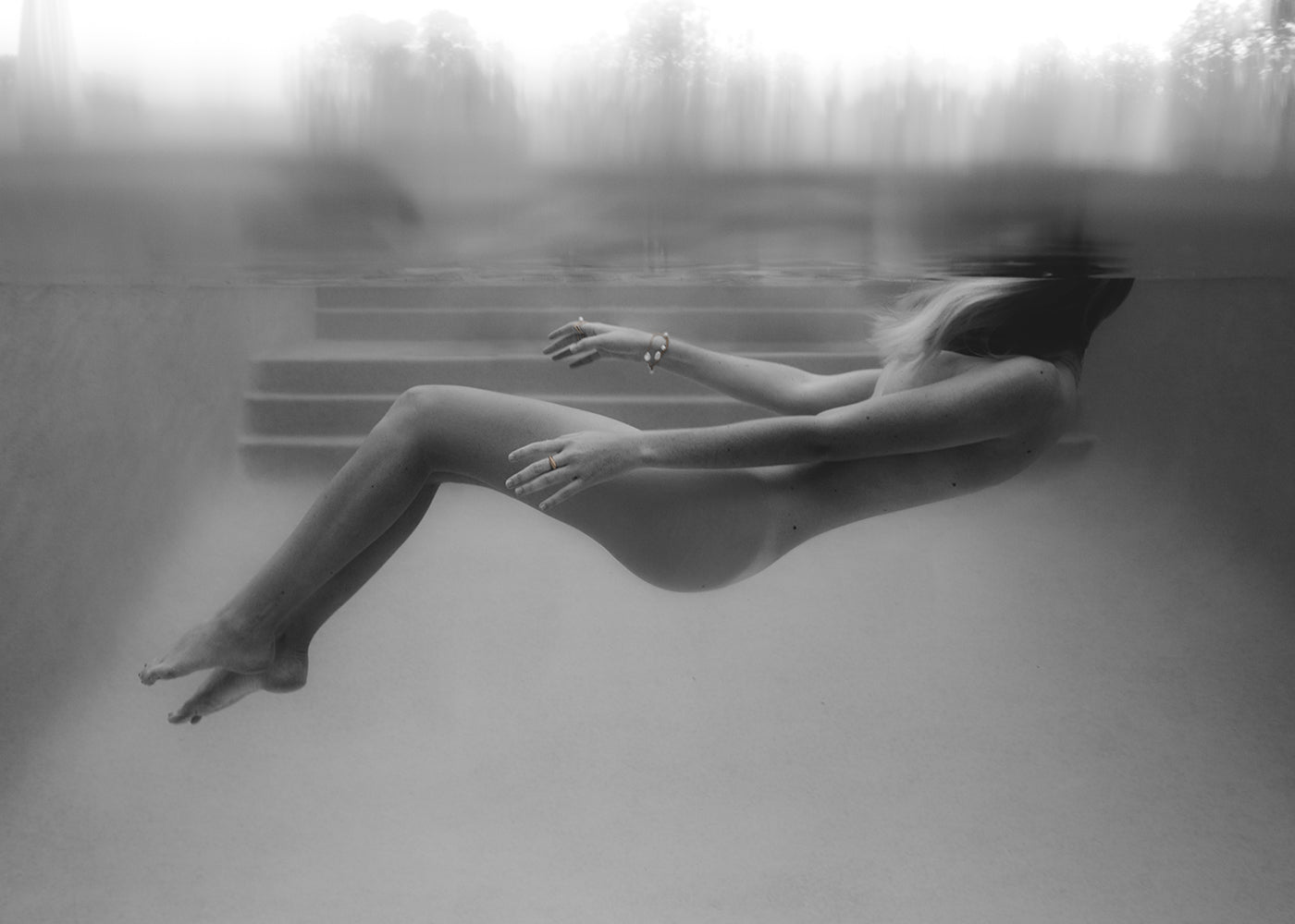 Francesca Owen's "Unveiled 04" captures a black and white image of a person underwater, seemingly floating just below the surface, facing upward with legs extended and arms slightly bent. The background features indistinct shapes above the waterline, hinting at an outdoor setting. This artwork is available as fine art prints, each accompanied by a certificate of authenticity.
