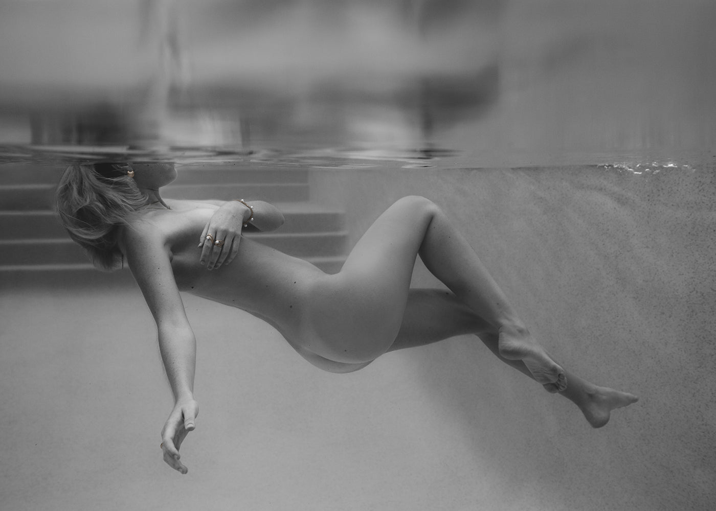 A person is floating underwater in a pool, partially submerged with their face obscured by the water's surface. The calm environment, highlighted by steps leading out of the pool in the background, captures a serene moment. This black and white image, titled "Unveiled 05" from Francesca Owen, exudes elegance and is perfect for fine art prints and custom framing.
