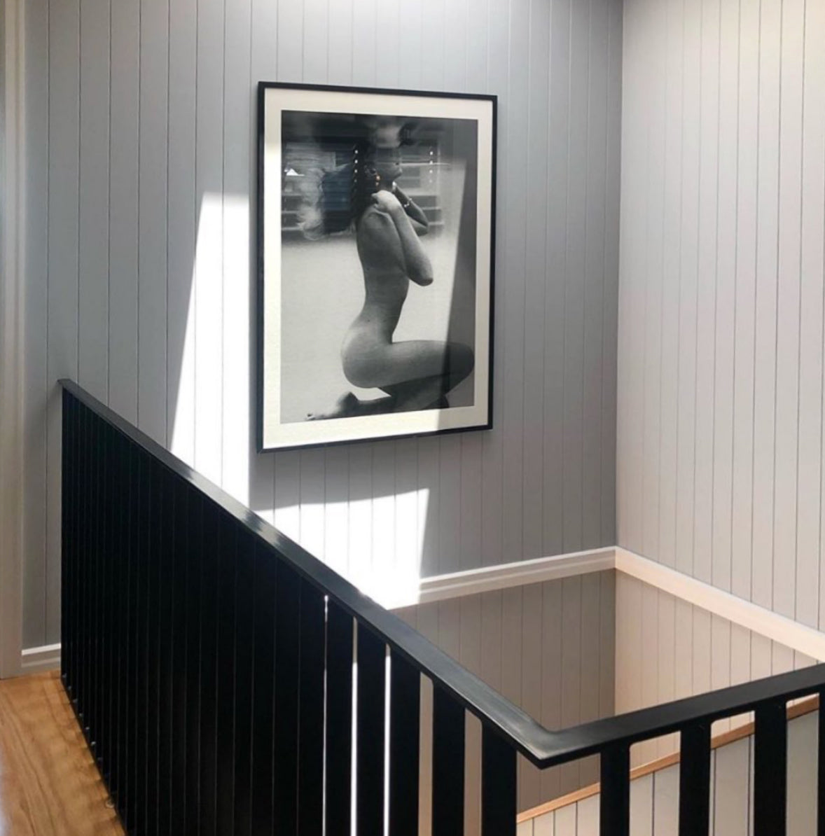 An artful black-and-white photograph from the "UNVEILED" collection by Francesca Owen, printed on fine art paper with archival pigments, graces a white shiplap wall. The image captures a partially clothed person in a seated partial profile. Below this framed Limited Edition print, a sofa adorned with grey and white cushions is partially visible.