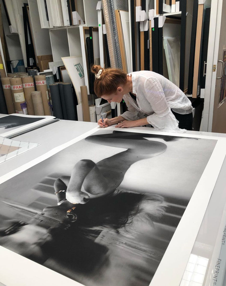 UNVEILED captures a serene black and white photograph by Francesca Owen, featuring a person underwater in a pool. The individual is kneeling with their head tilted back, hair floating, and reflections on the water surface creating a mirrored effect. Adorned with visible gold earrings and bracelet, this limited edition piece is printed on fine art paper using archival pigments.